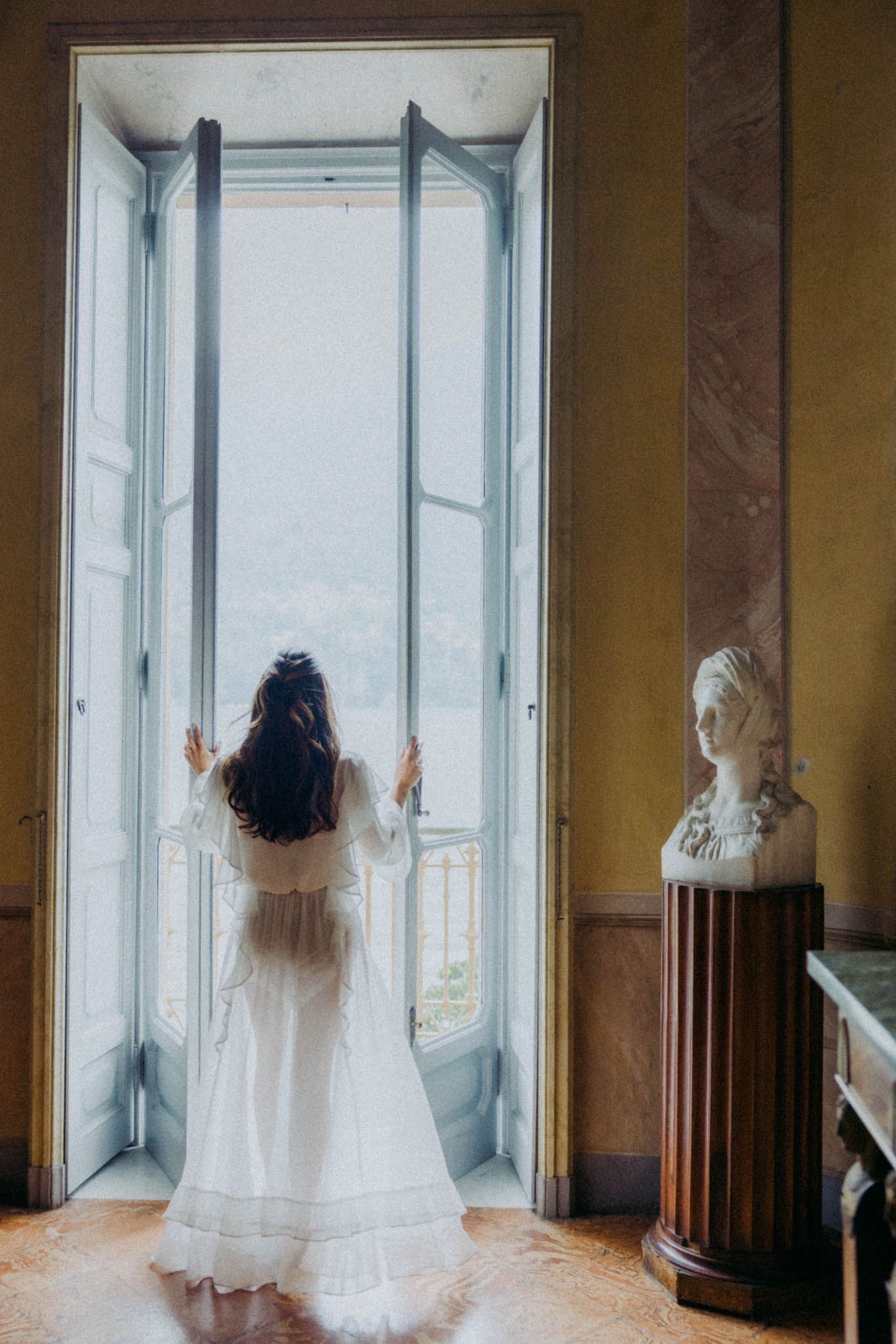 A flowing white dress photographed from behind against large windows in an elegant interior.