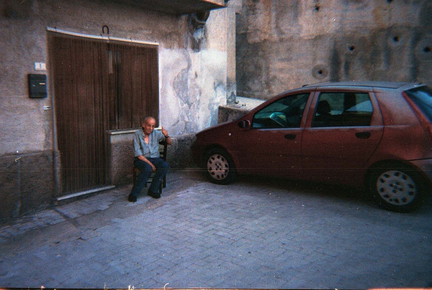 A vintage photo shows a parked red car next to a weathered building wall with a wooden door.