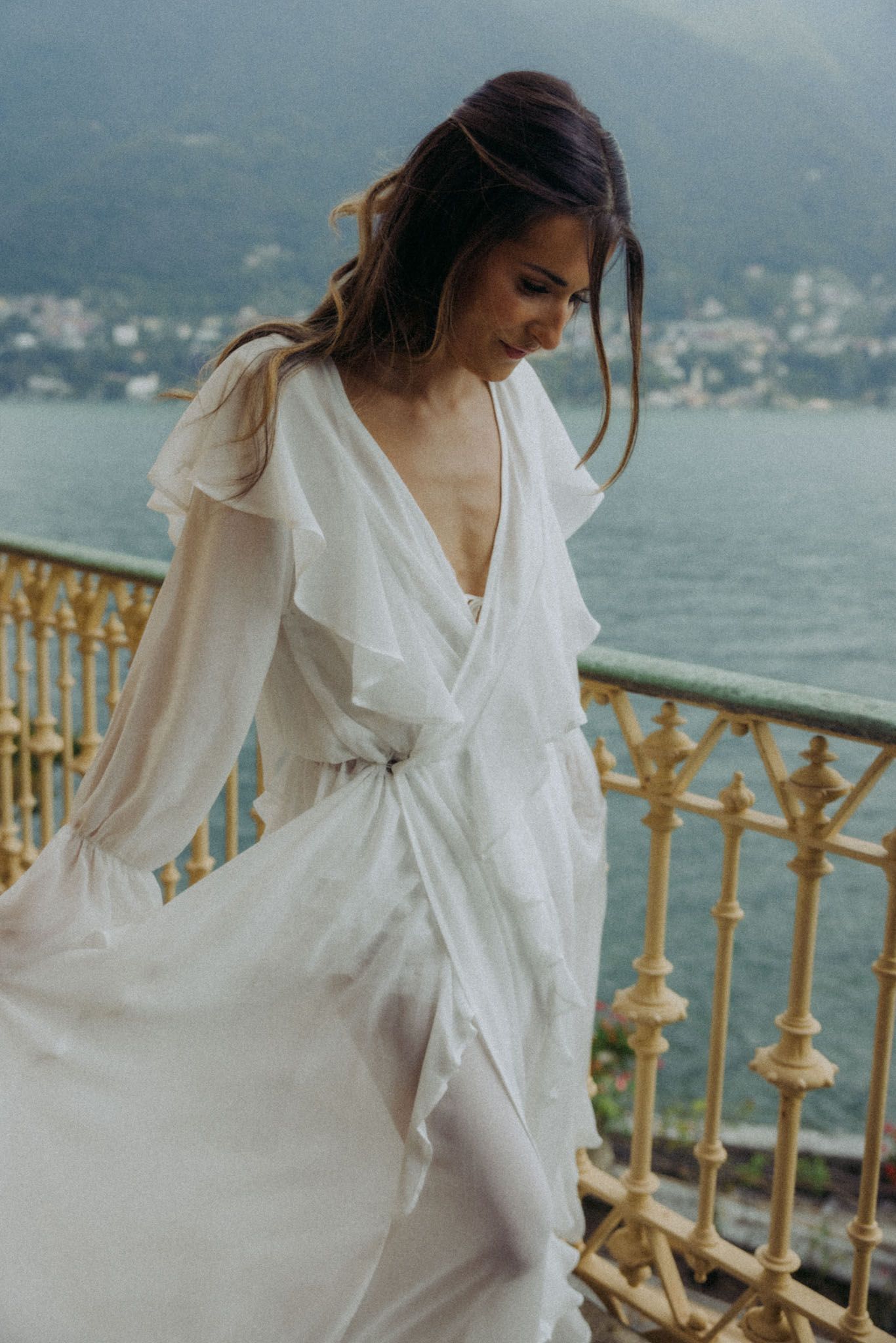 A bride in a flowing white dress poses on an ornate balcony overlooking a scenic lake and mountains.