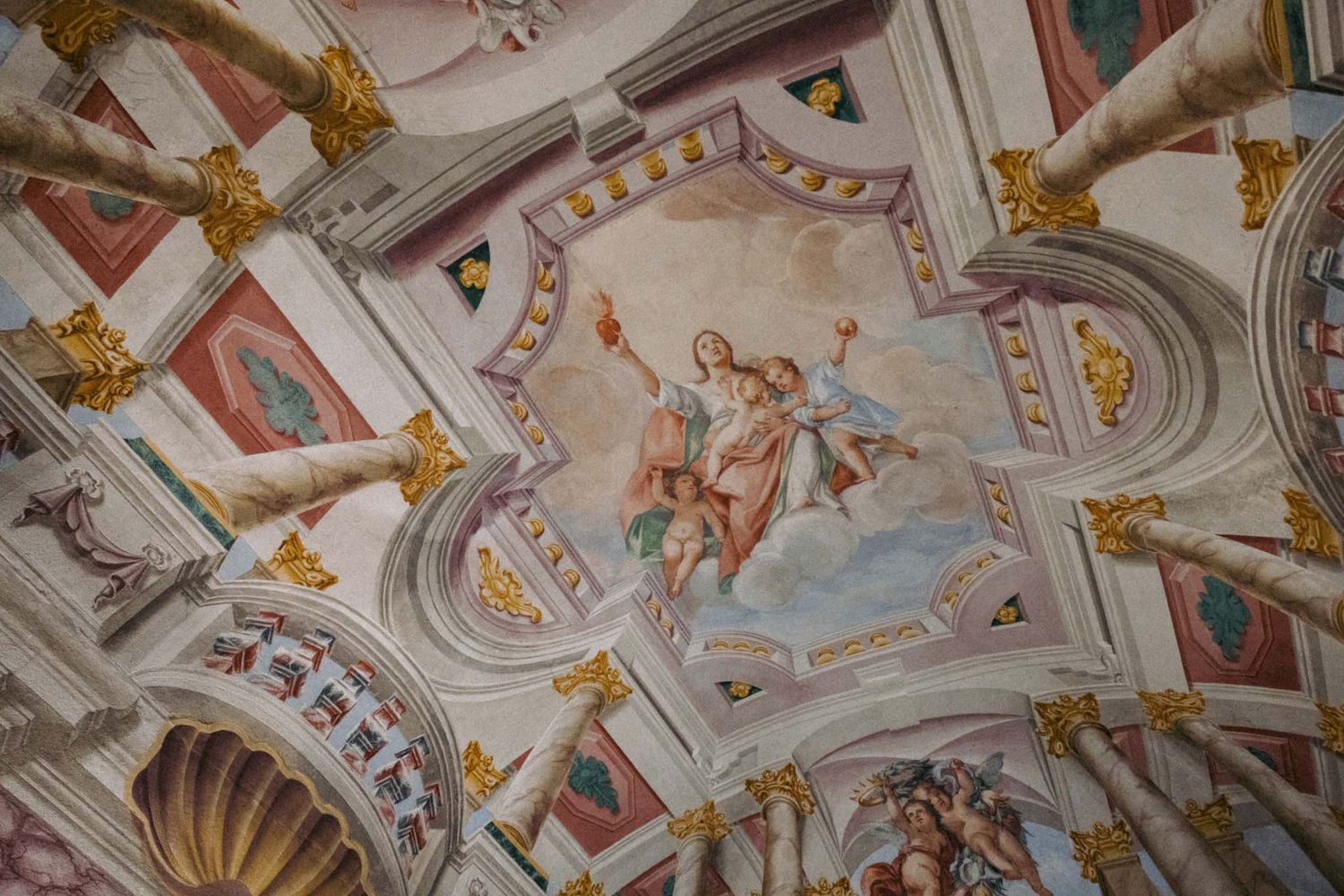 Ornate baroque church ceiling with intricate frescoes, gilded decorations, and painted religious scenes in vibrant colors.