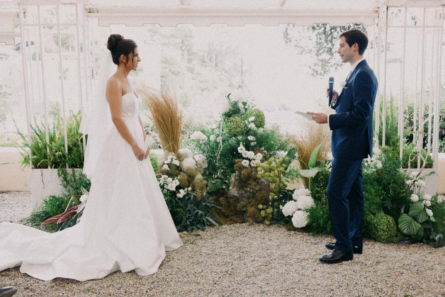 A couple in wedding attire walks along a garden path during their outdoor ceremony.