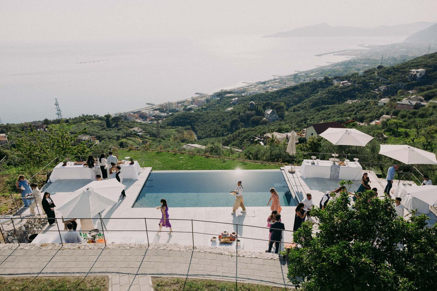 Panoramic view of a white infinity pool overlooking a scenic mountainous coastal landscape on a sunny day.