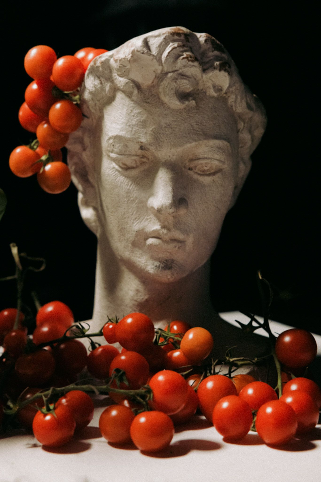 A classical marble bust sculpture surrounded by fresh red cherry tomatoes on the vine against a dark background.
