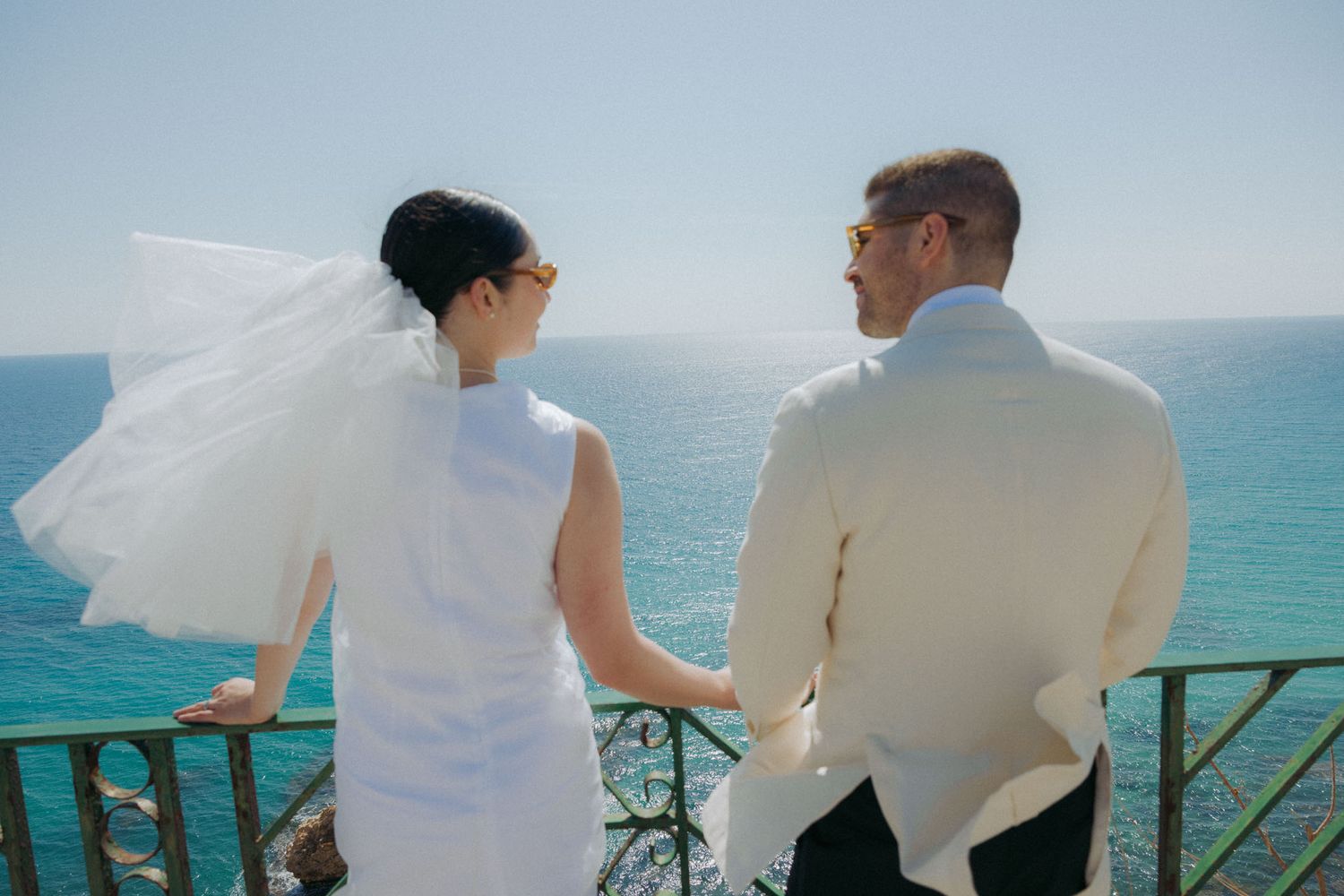 People dressed in white outfits sitting and chatting on a terrace overlooking turquoise ocean waters.