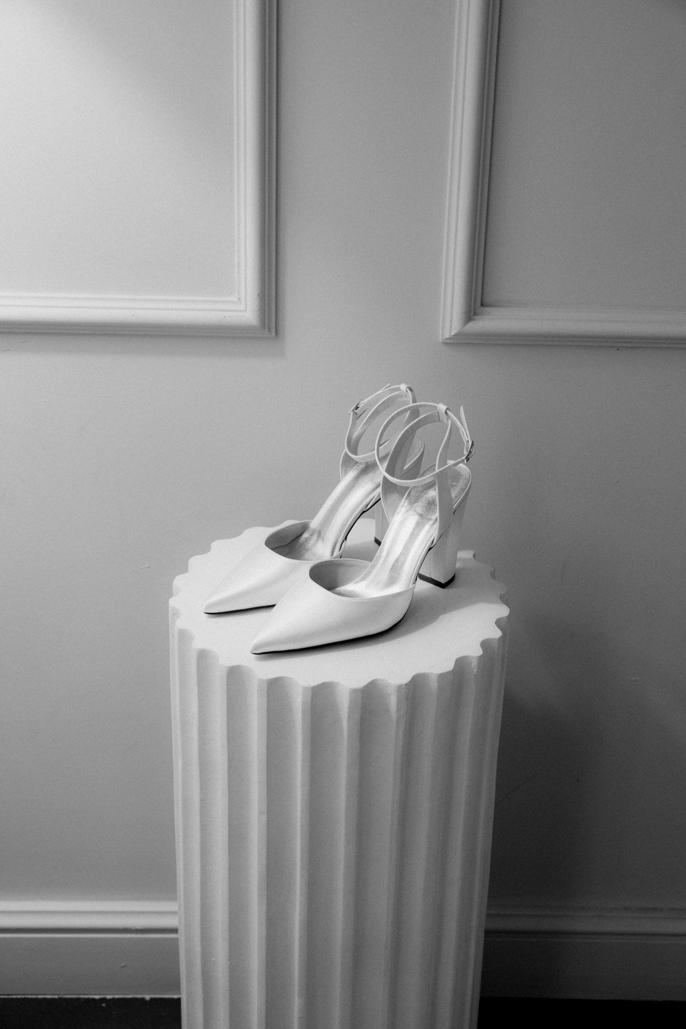 A white high-heeled shoe sits elegantly atop a fluted pedestal against a light-colored wall in black and white.