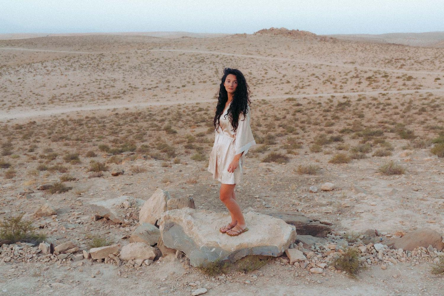 A person in a white dress stands on a large rock in a vast desert landscape during golden hour.