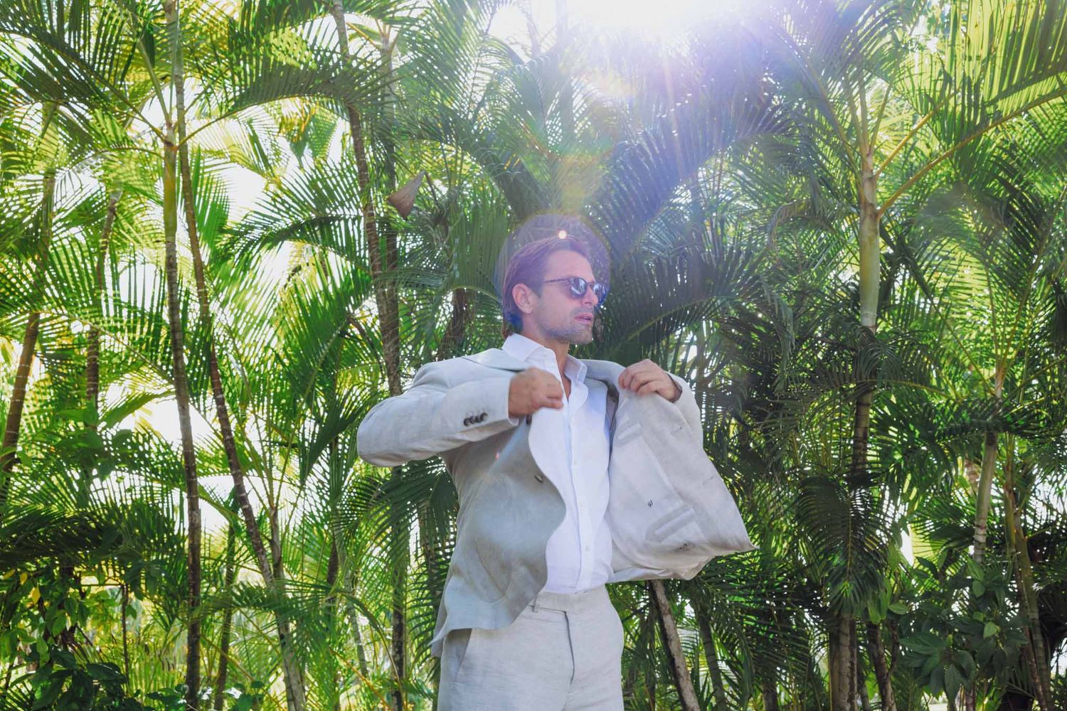 A person in white clothing dances joyfully among lush tropical palm trees on a sunny day.