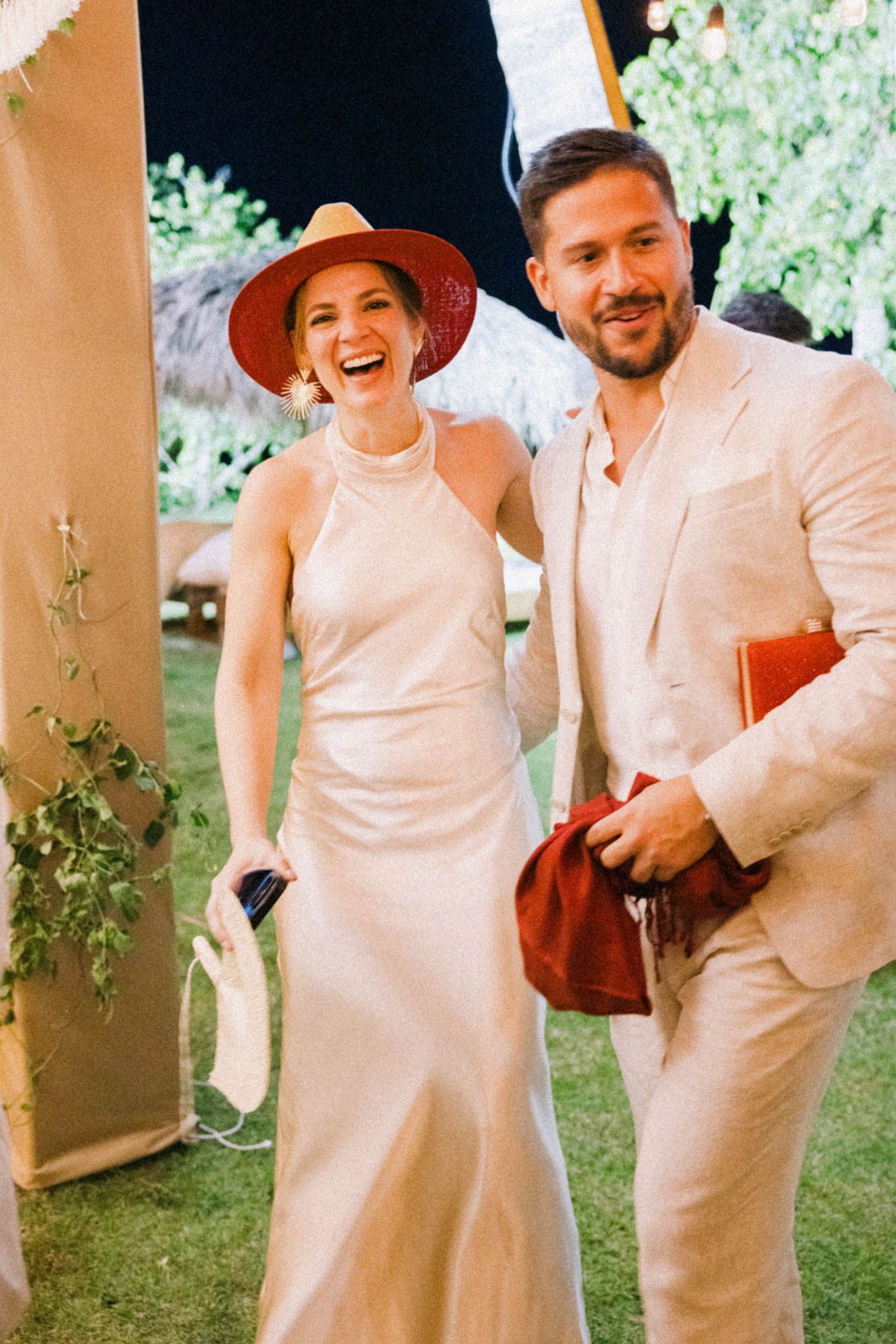 A couple in matching white outfits poses playfully in a garden setting with stone columns and greenery.