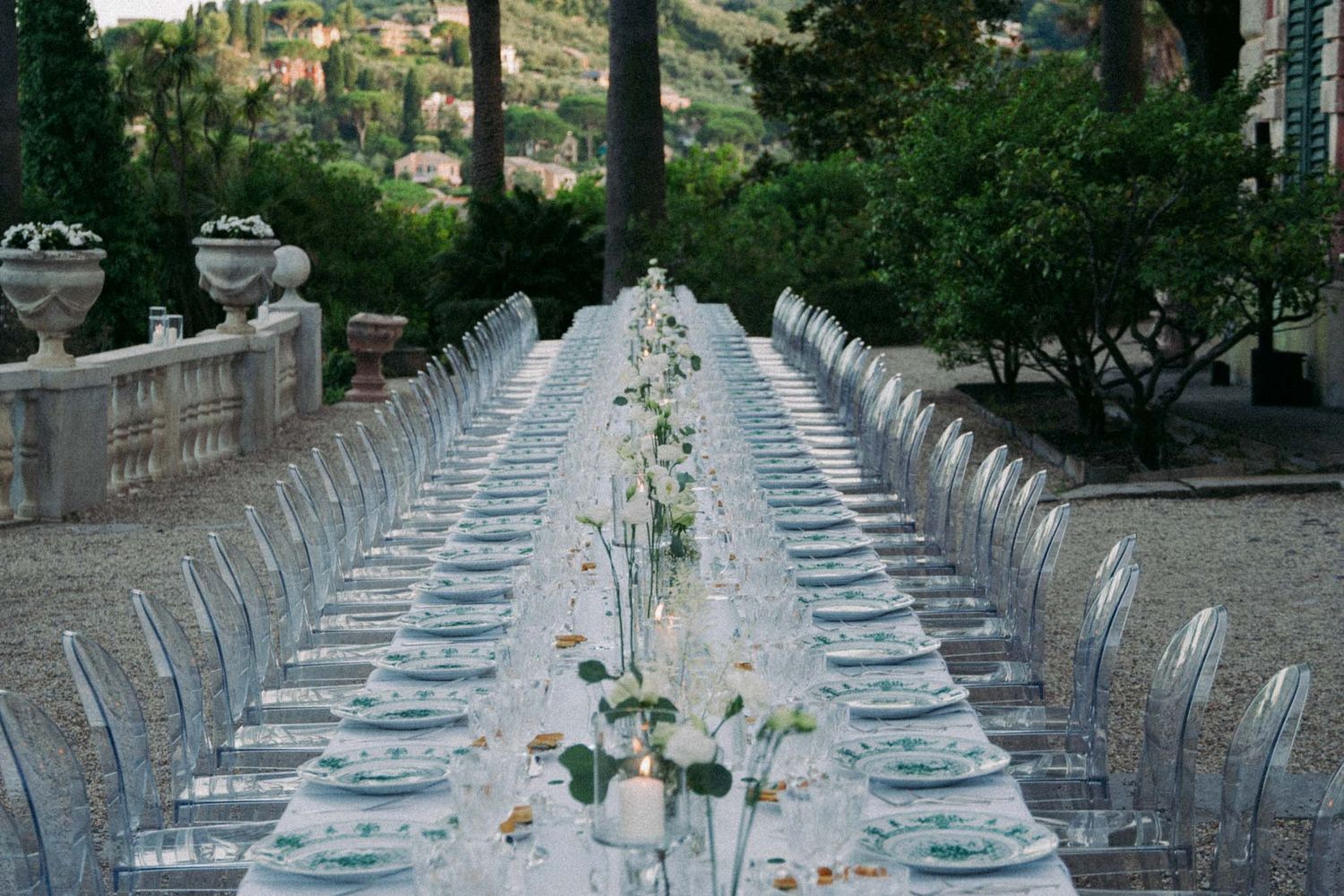 Long elegant outdoor dining table set with white linens and ghost chairs stretches along a stone terrace.