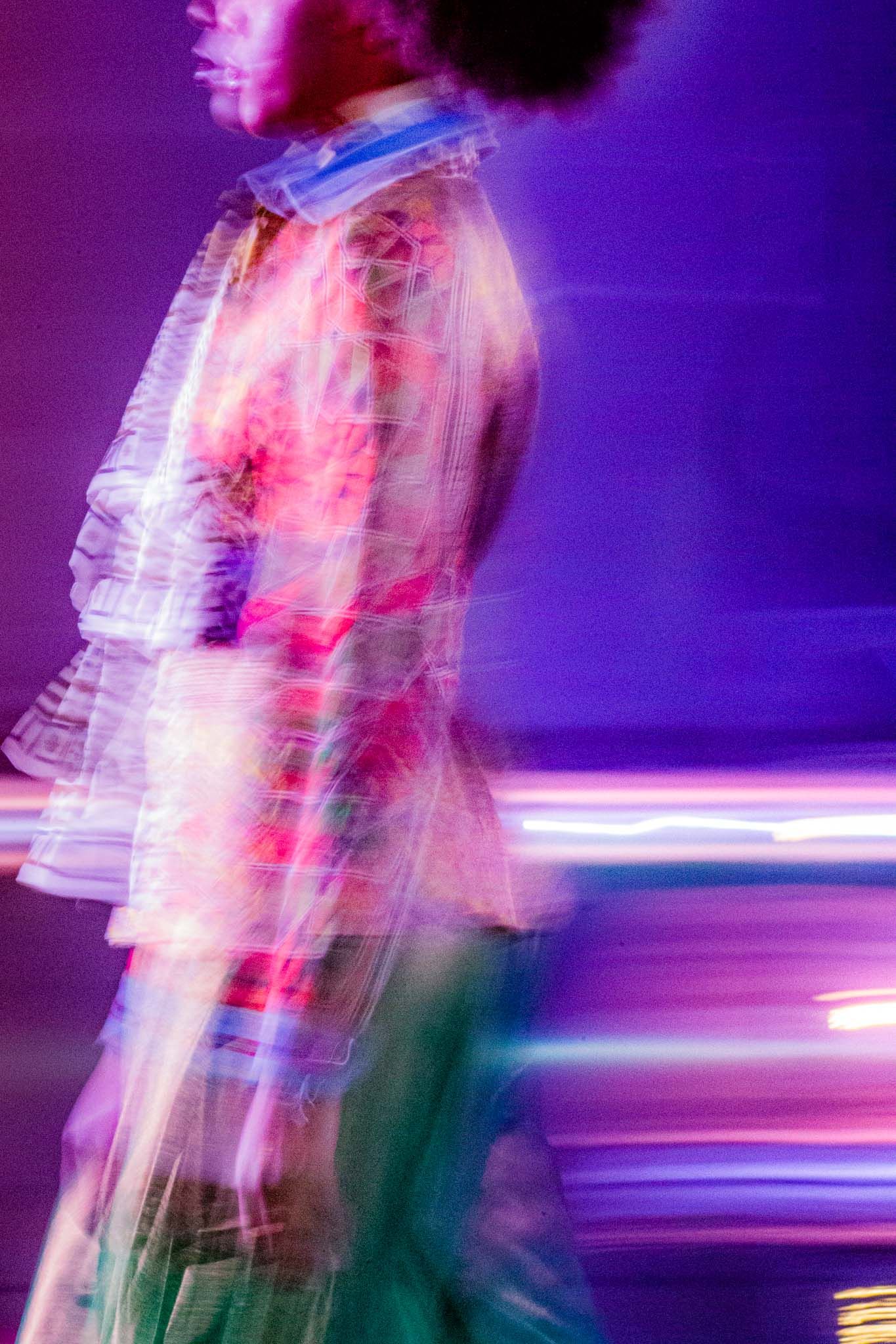 Long exposure photo shows blurred motion of a person in a dress creating colorful light trails against a purple background.