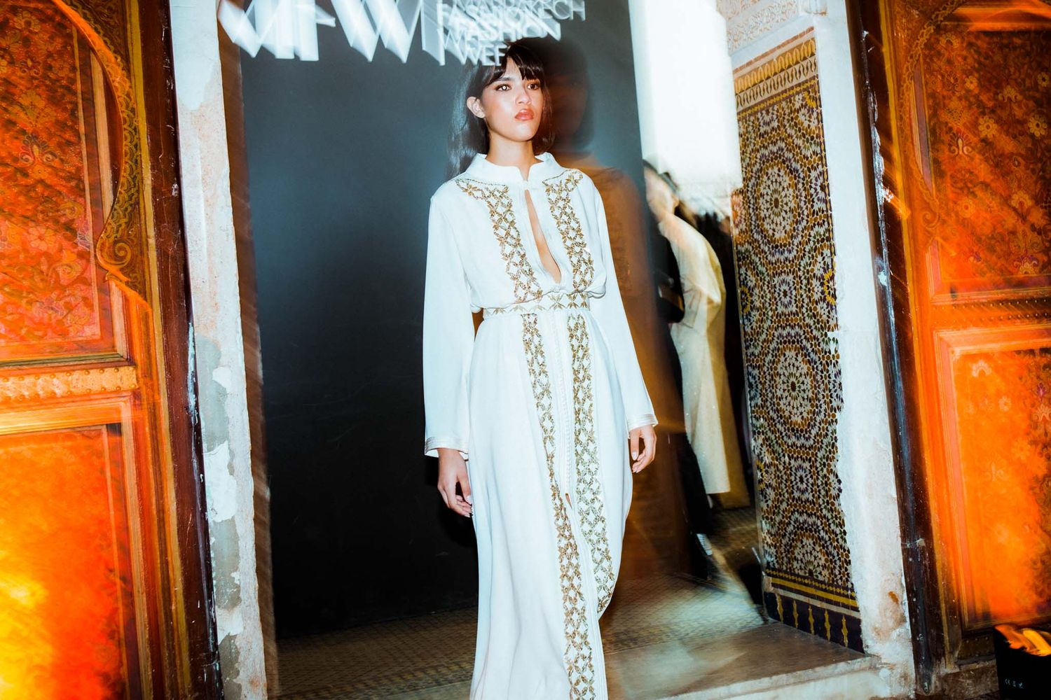 A person wearing a white lace dress stands in an ornate hallway with warm orange lighting and decorative walls.