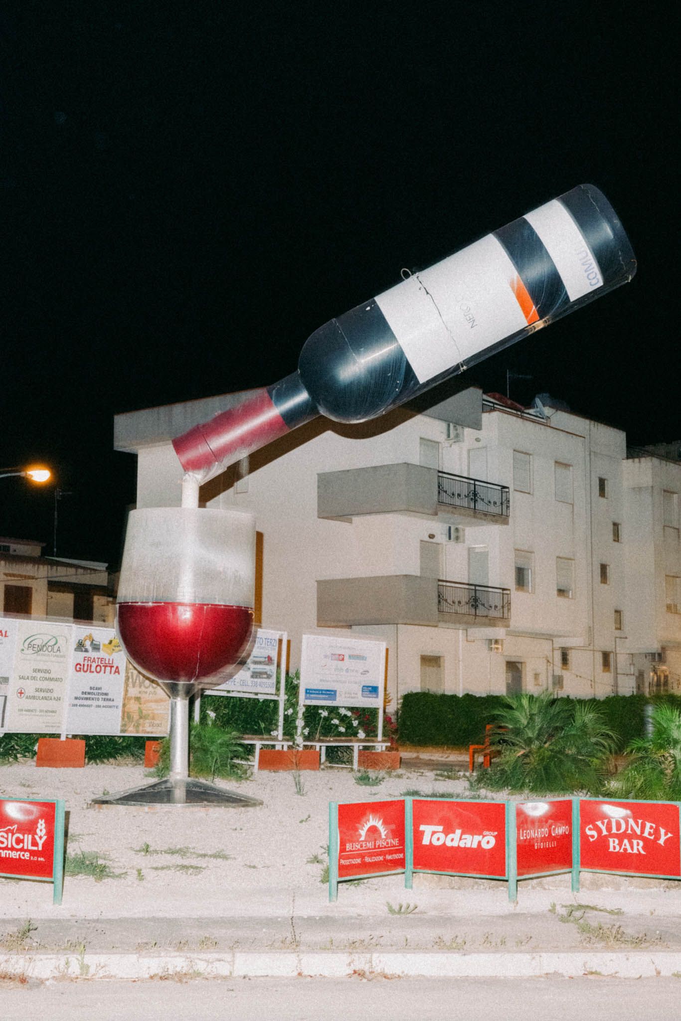 Giant wine bottle and glass sculptures displayed along a white wall at night with illuminated buildings in the background.