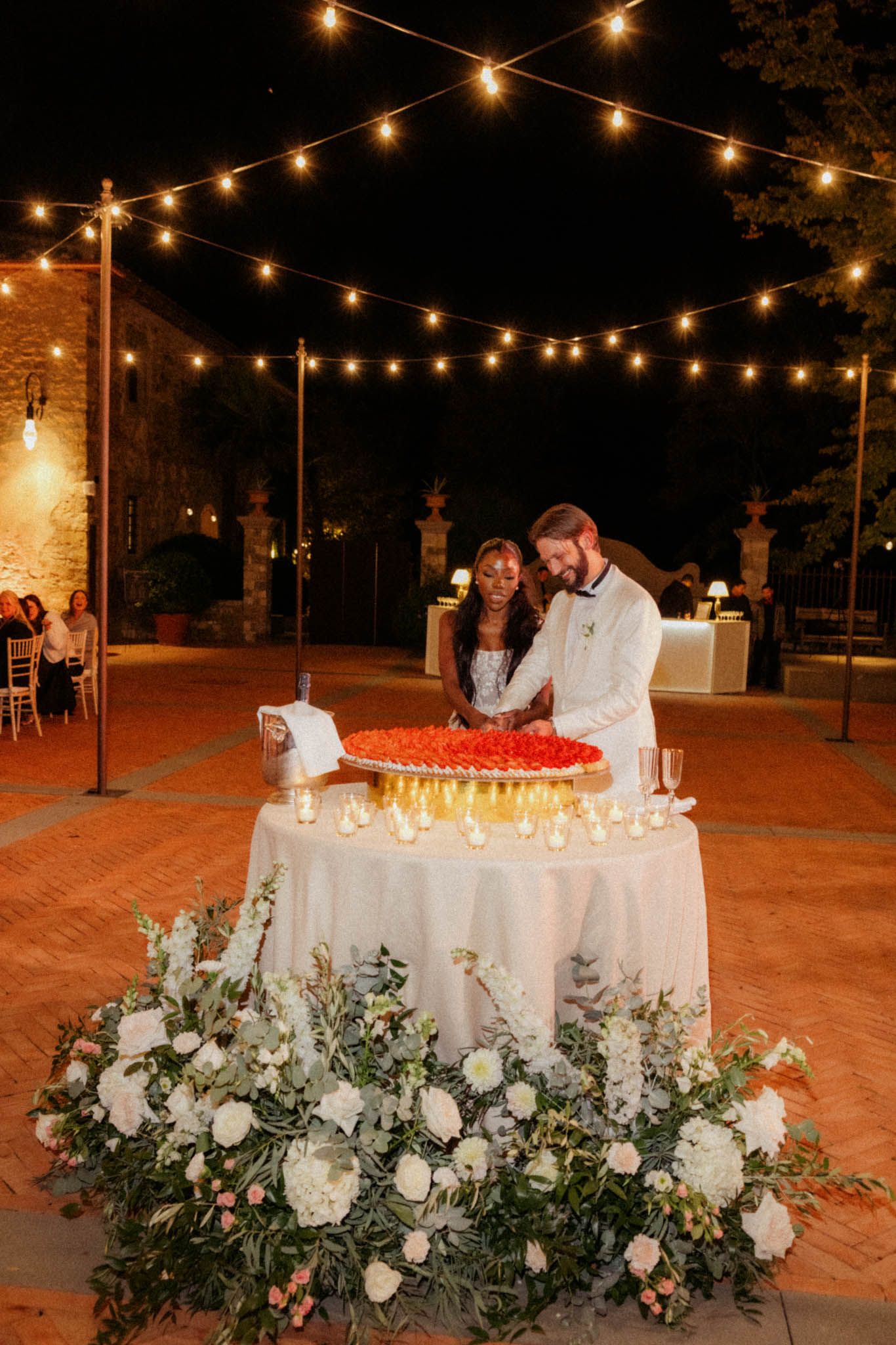Elegant wedding cake display with string lights and floral arrangements at an outdoor evening reception.