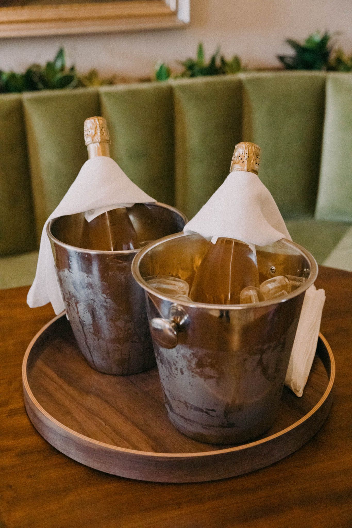 Two champagne bottles chilling in ice buckets with white napkins on a wooden tray against green booth seating.