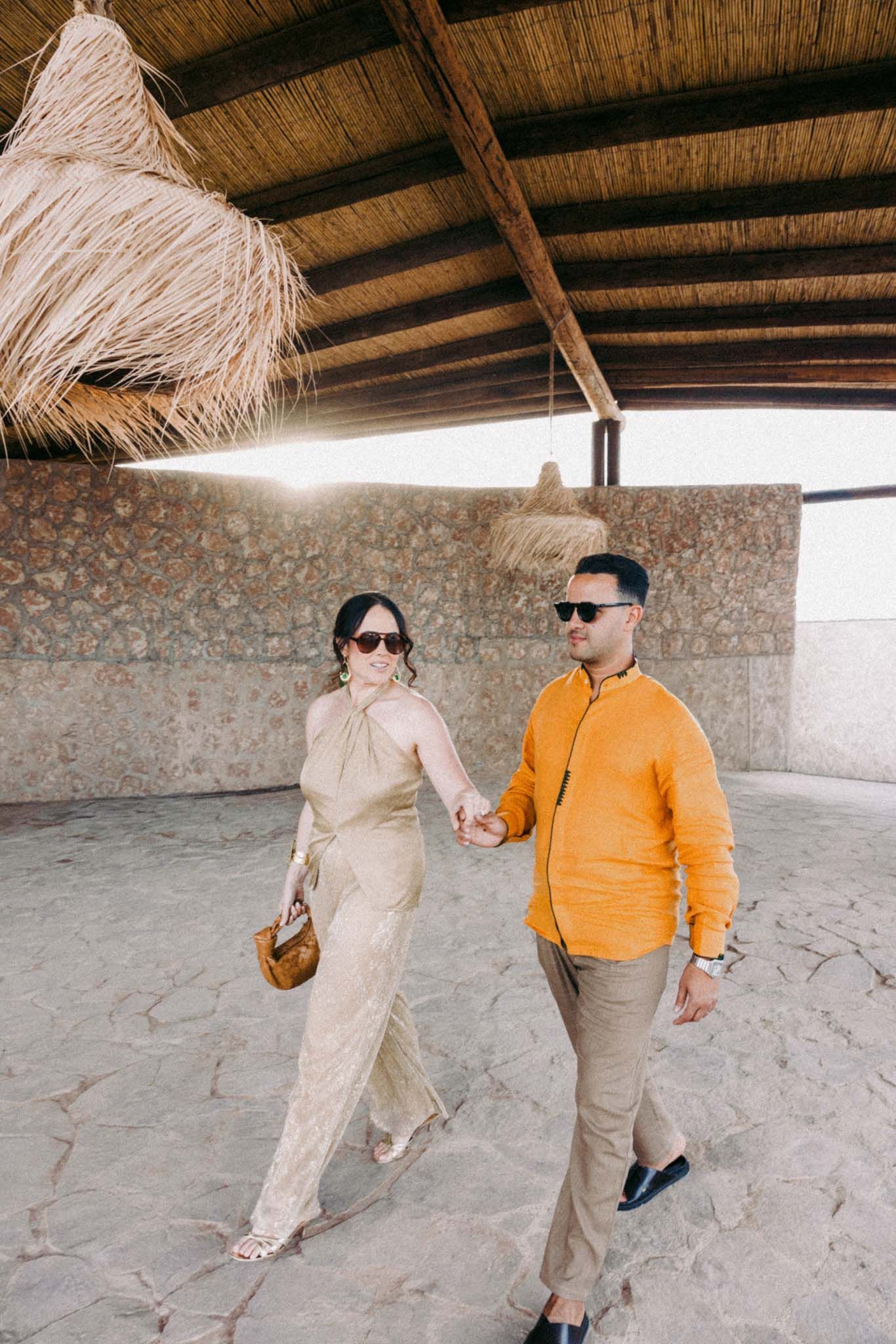 Two people in casual attire walking together under a rustic thatched roof structure with stone walls.