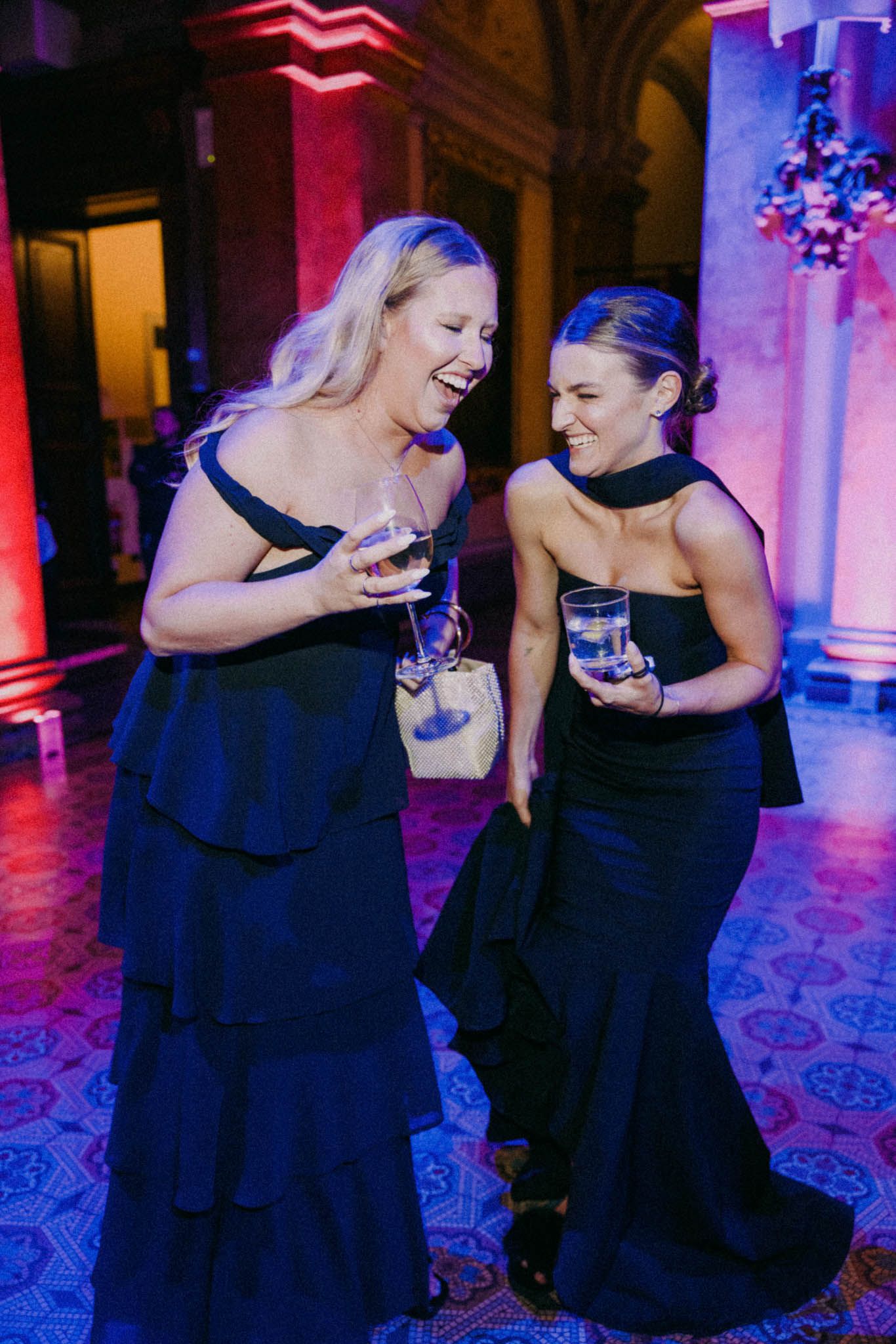 Two people in black dresses share a joyful moment on the dance floor illuminated by colorful purple and pink lighting.