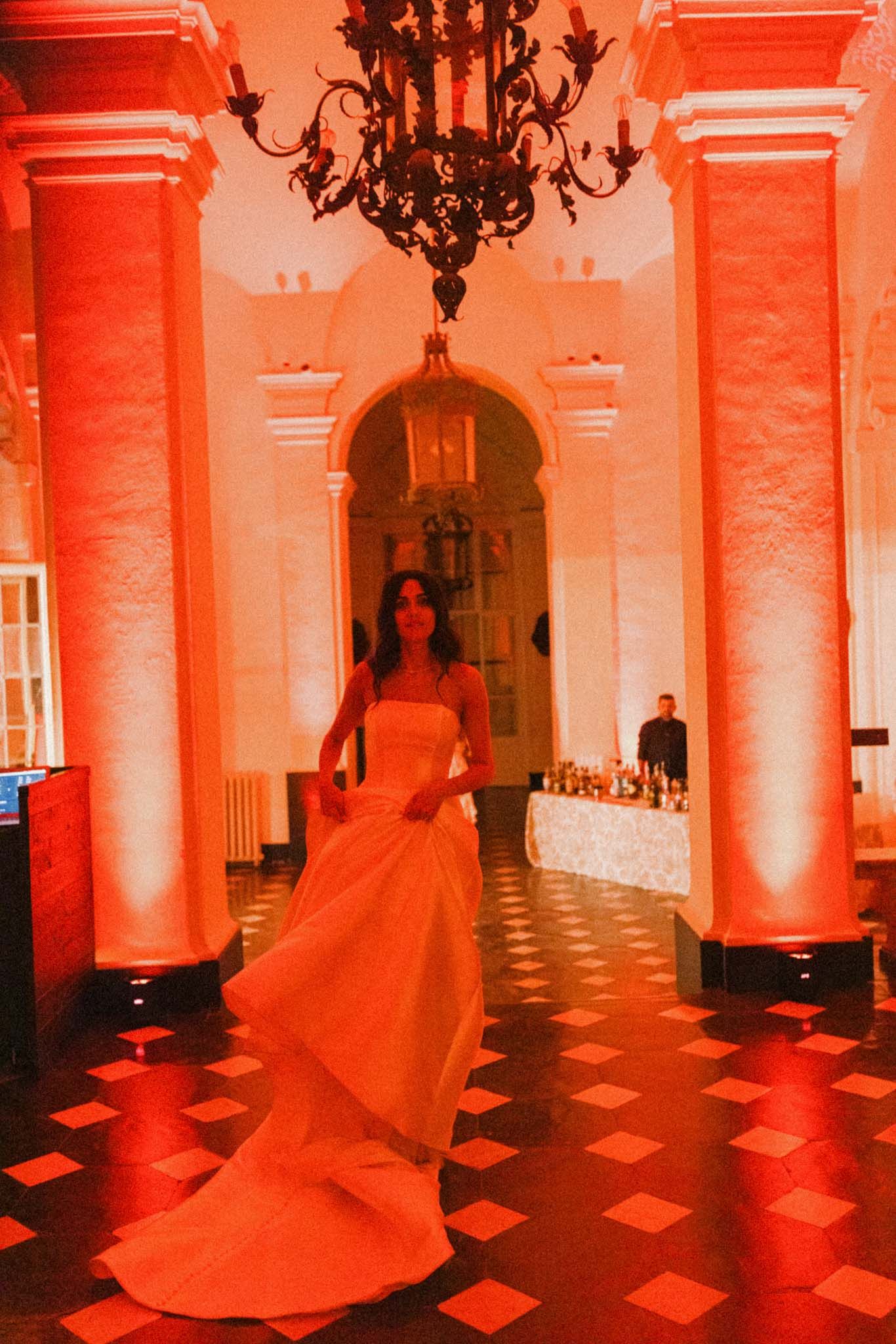 A figure in a flowing red gown twirls across a checkered floor in a grand columned hall bathed in dramatic red lighting.