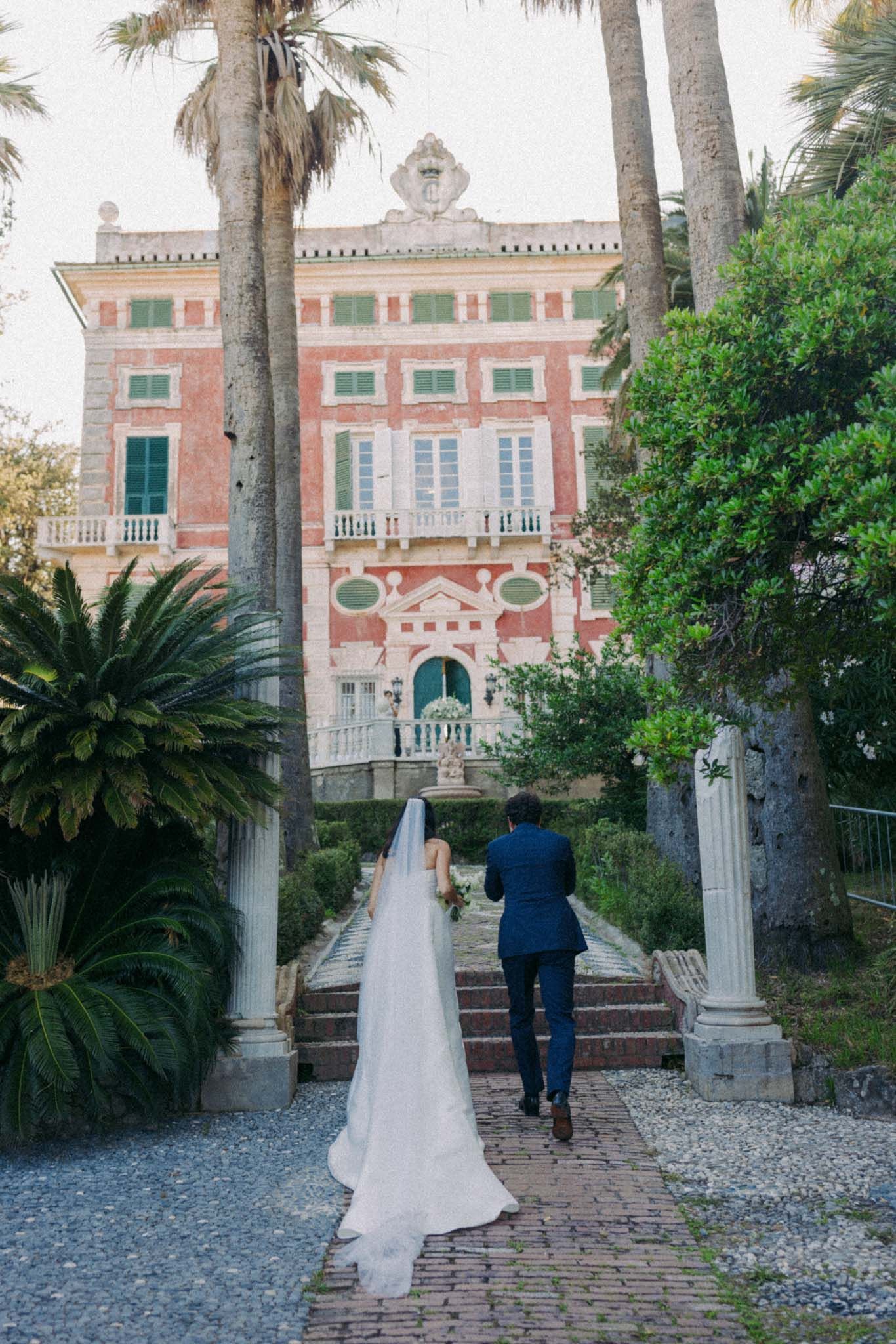 A wedding couple walks along a brick path towards a pink Mediterranean-style villa surrounded by palm trees.