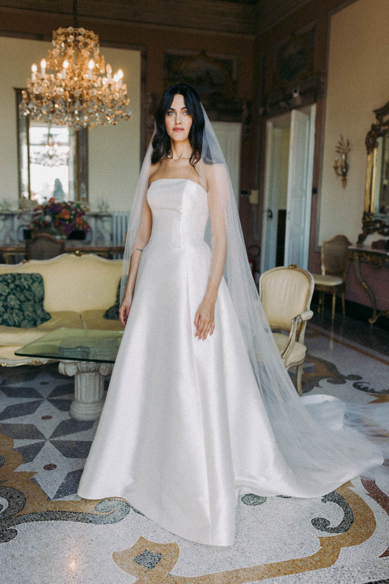 Elegant strapless white wedding gown with long veil photographed in luxurious room with crystal chandelier and ornate floor.