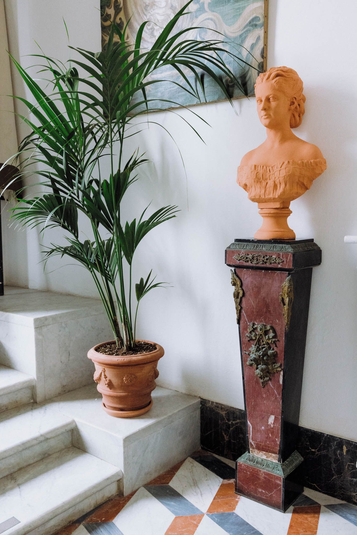 A classical interior scene with terracotta busts and potted palm plants along white stairs and tiled floors.