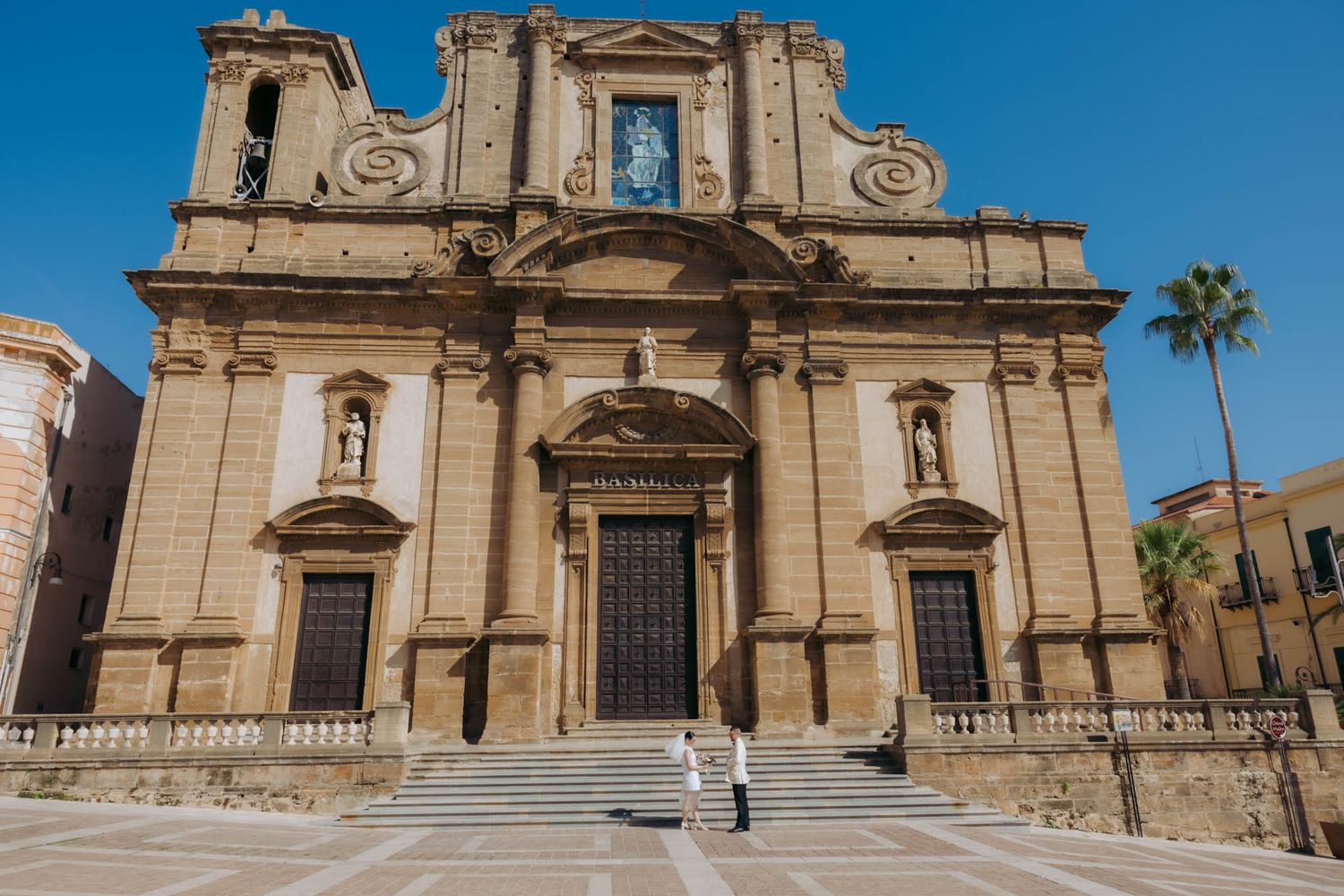 Historic baroque church facade with twin towers and stone steps in Agrigento, Sicily against bright blue sky and palm tree.