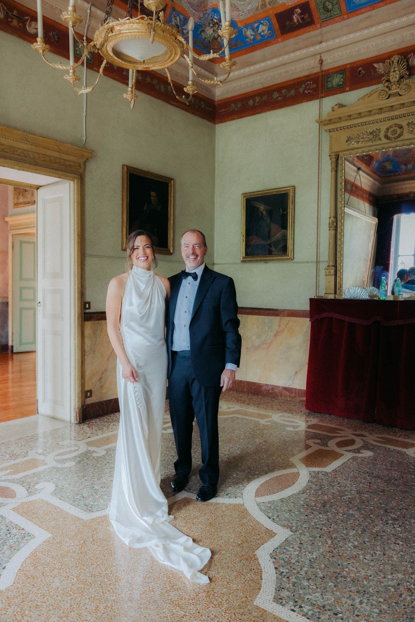 Two people stand in an ornate historic room with decorative ceiling frescoes and patterned marble floors.