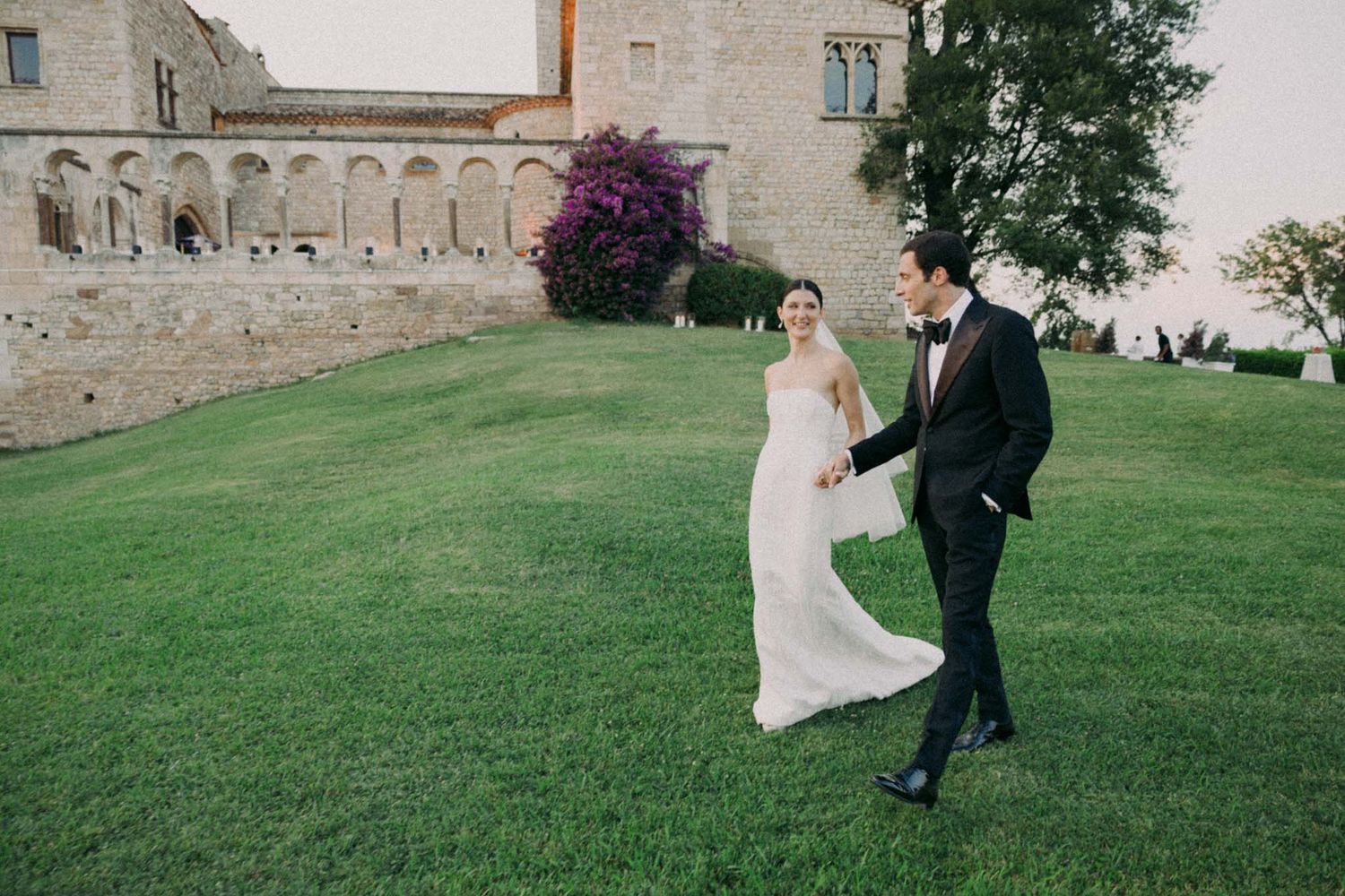 A couple in formal wedding attire shares a romantic dance together on a lush green lawn outside a stone building.