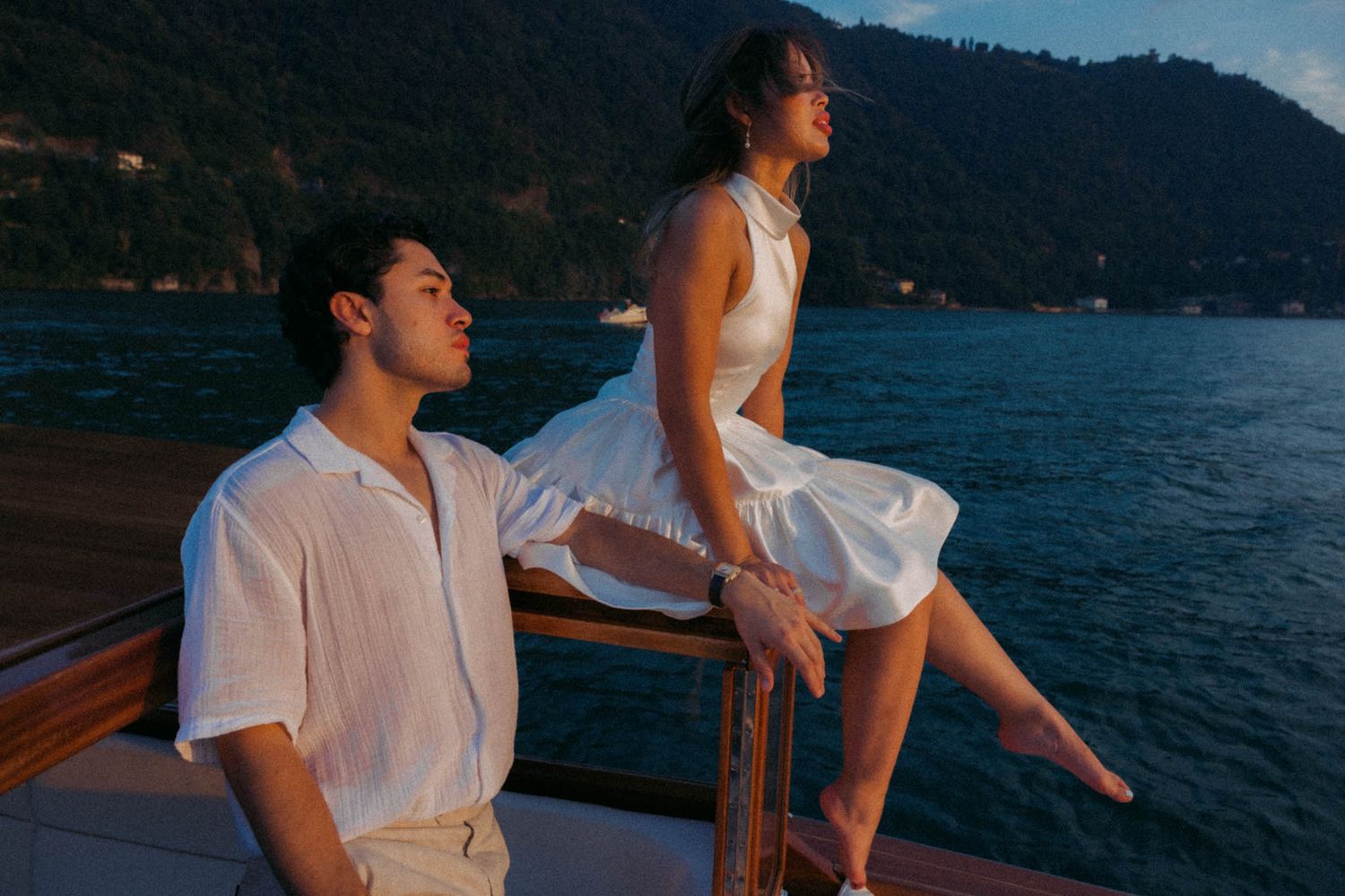 A couple sits on a wooden dock overlooking a serene lake surrounded by mountains at sunset.