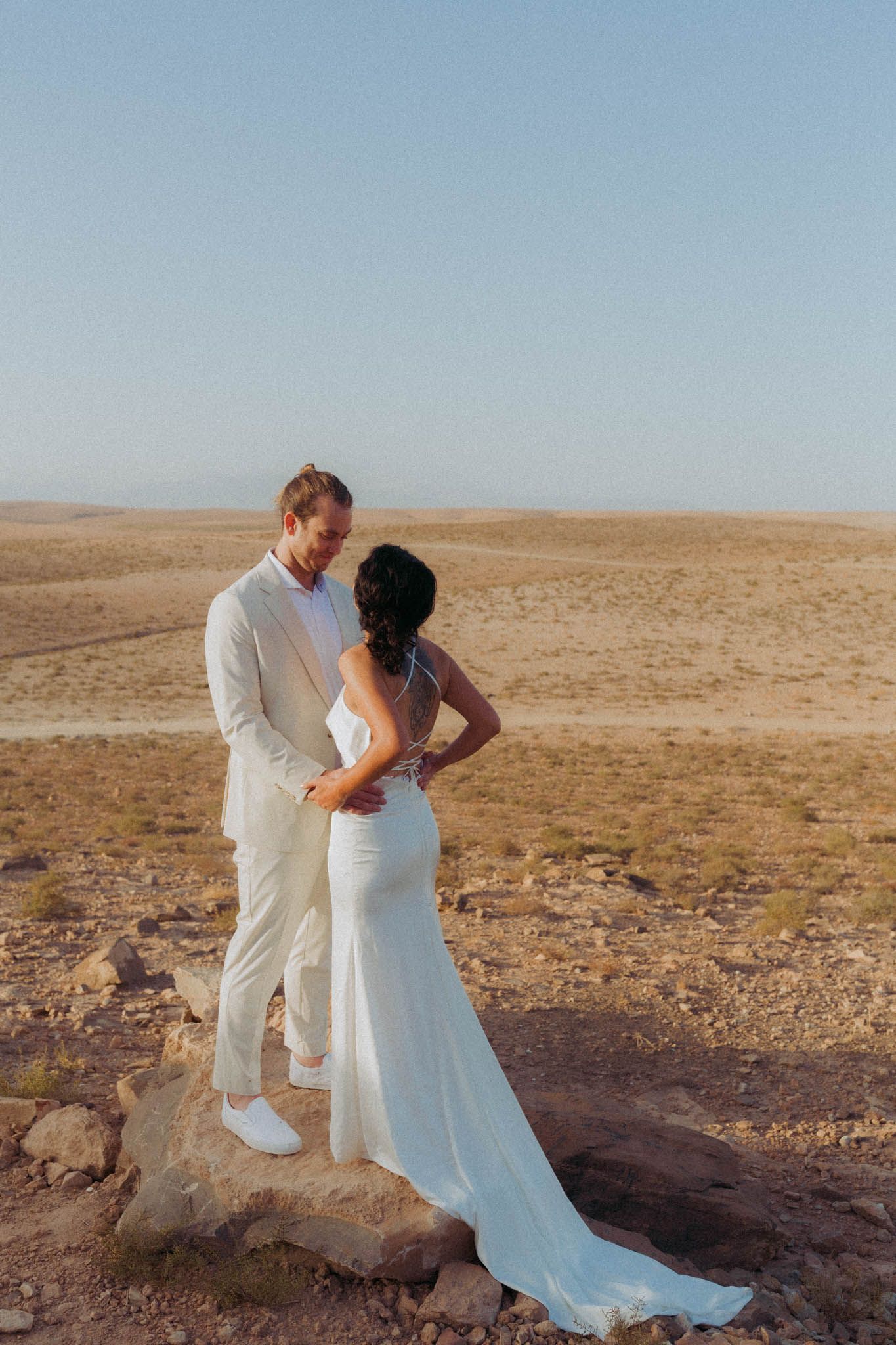 A couple in white wedding attire share romantic moments against a desert landscape backdrop.