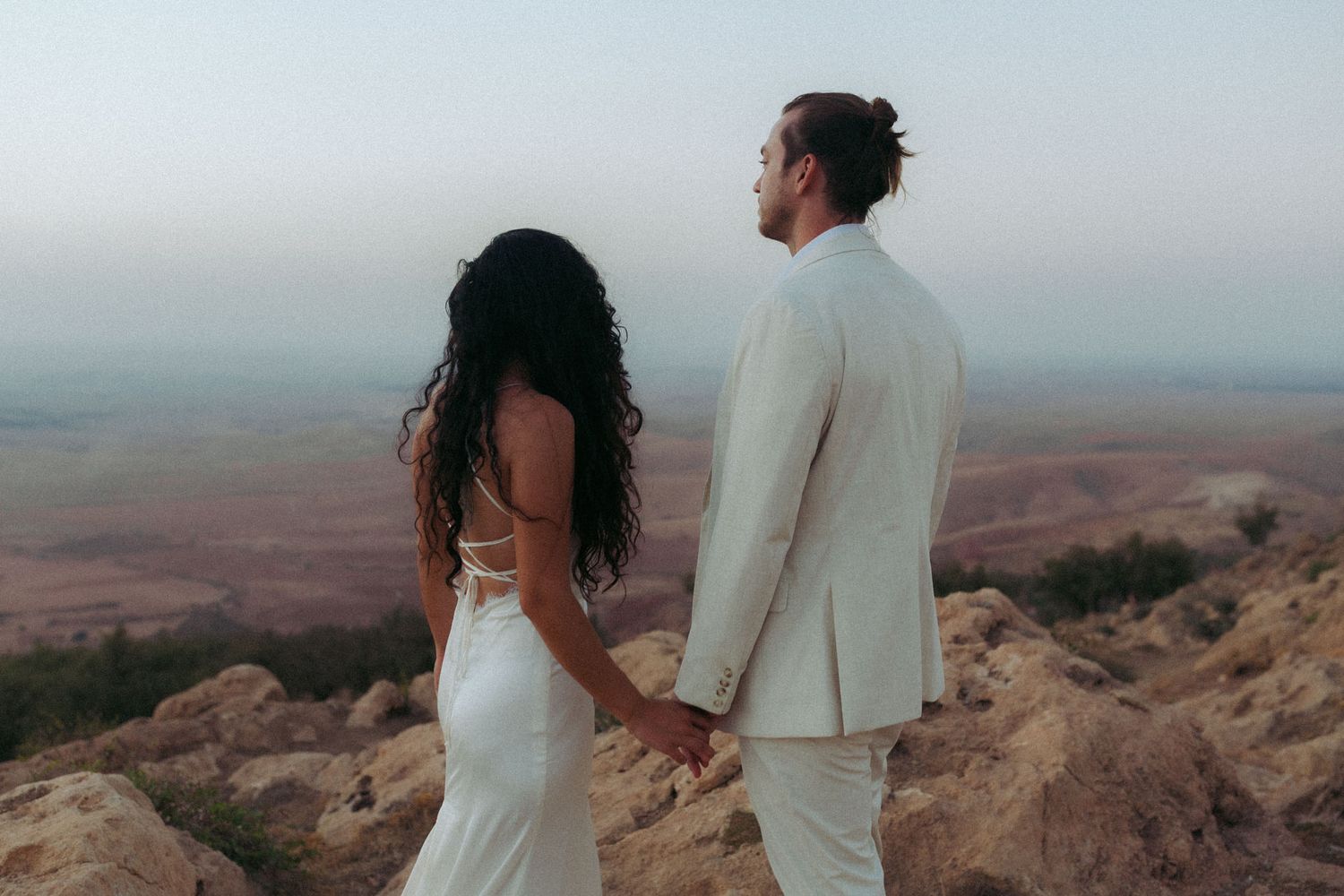 Couple in wedding attire stands together overlooking a scenic desert mountain landscape at sunset.