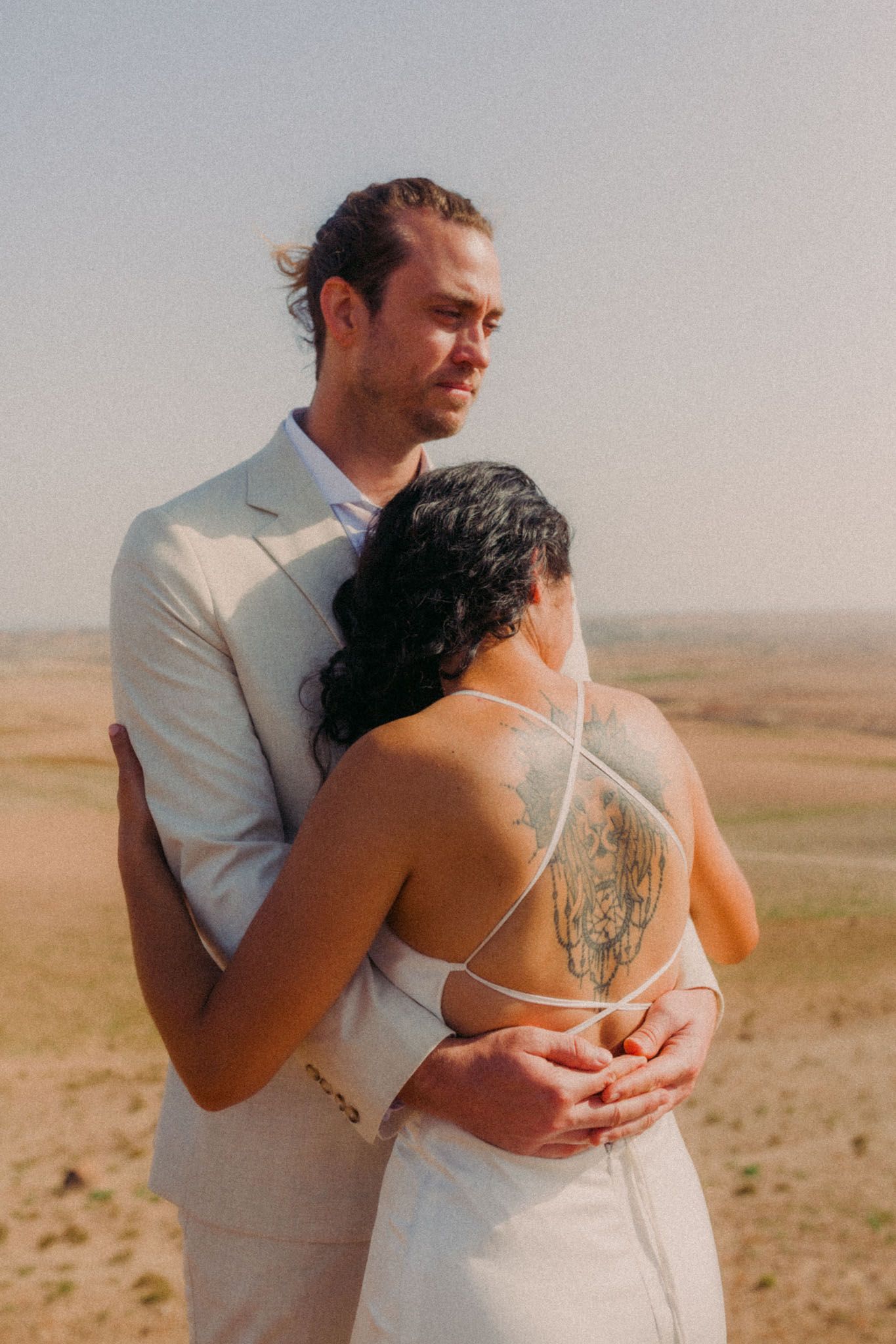 A couple embraces in an intimate moment against a desert landscape backdrop during a wedding photoshoot.