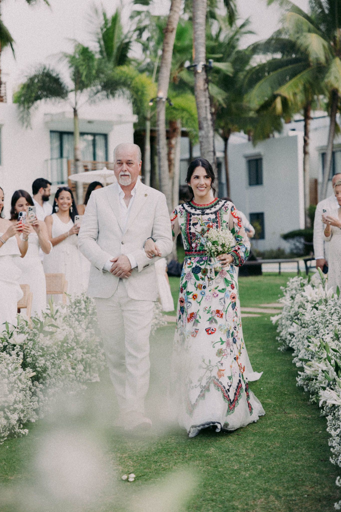 Wedding party walks along palm-lined garden path in white attire with floral accents.