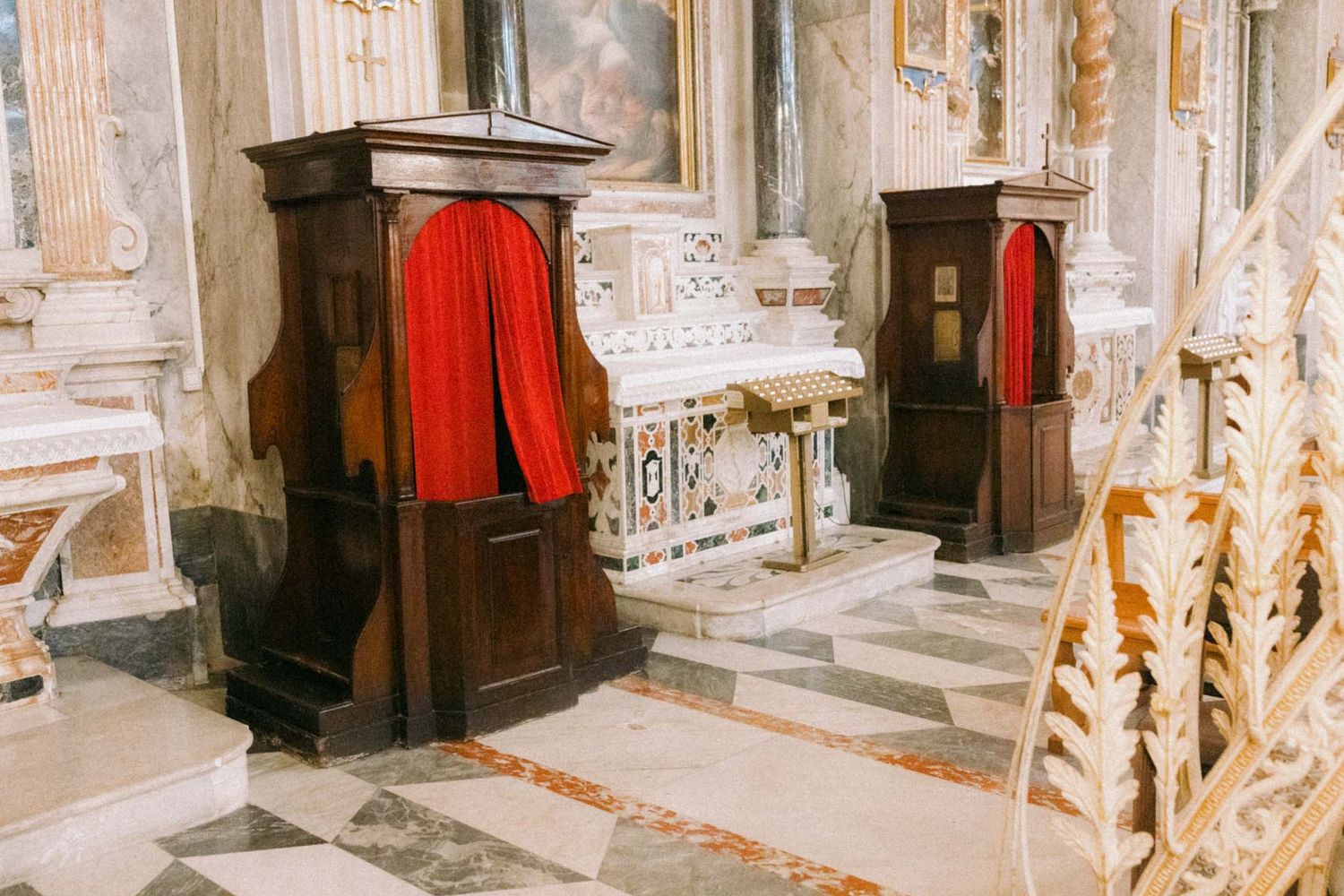 Two wooden confessional booths with red curtains inside an ornate marble church interior.