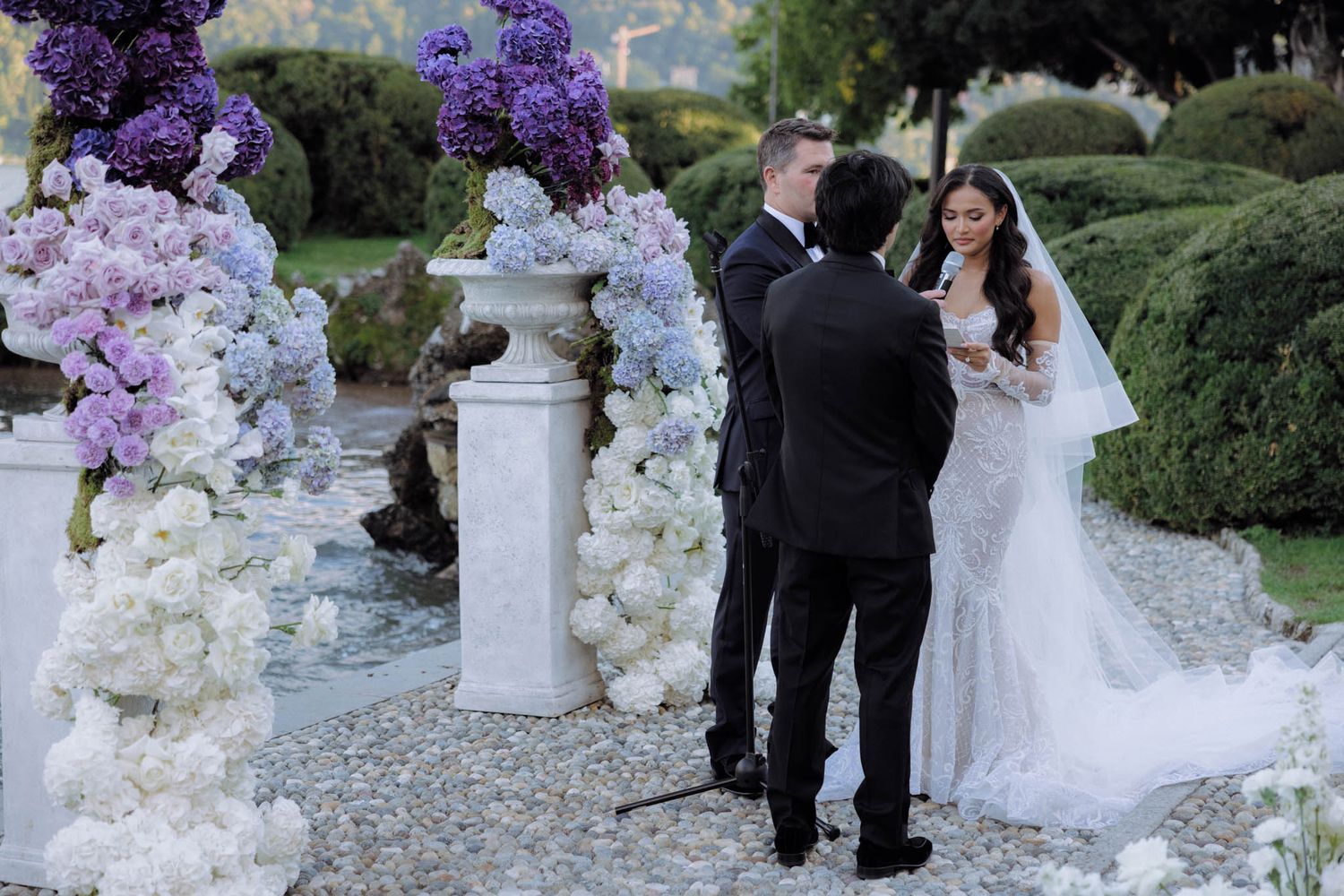 Wedding ceremony setup with purple and white floral arrangements on pedestals in an outdoor garden setting.