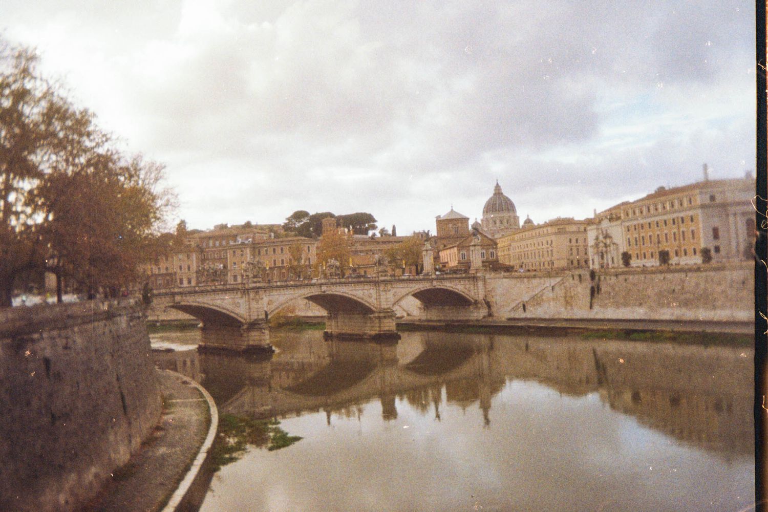 Historic stone buildings and bridges line a tranquil river waterway in Florence, Italy, reflecting in the calm waters.