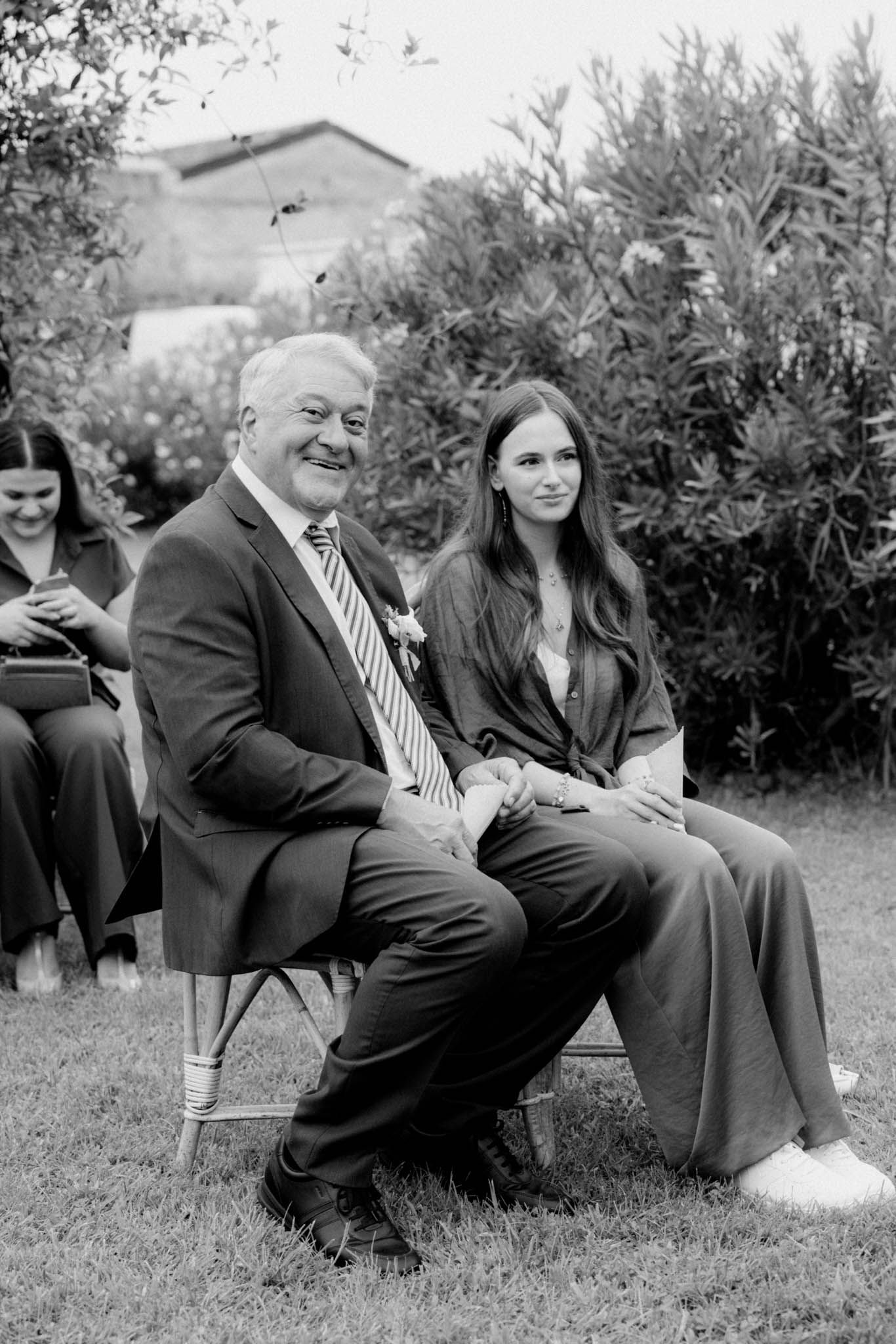 Black and white photo of people sitting outdoors on chairs in a garden setting during what appears to be a social gathering.