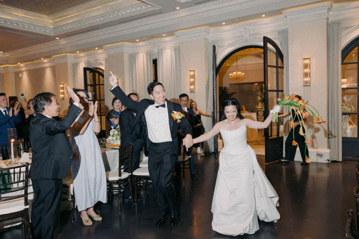 Wedding guests dance and celebrate during a reception in an elegant ballroom with ornate columns and archways.