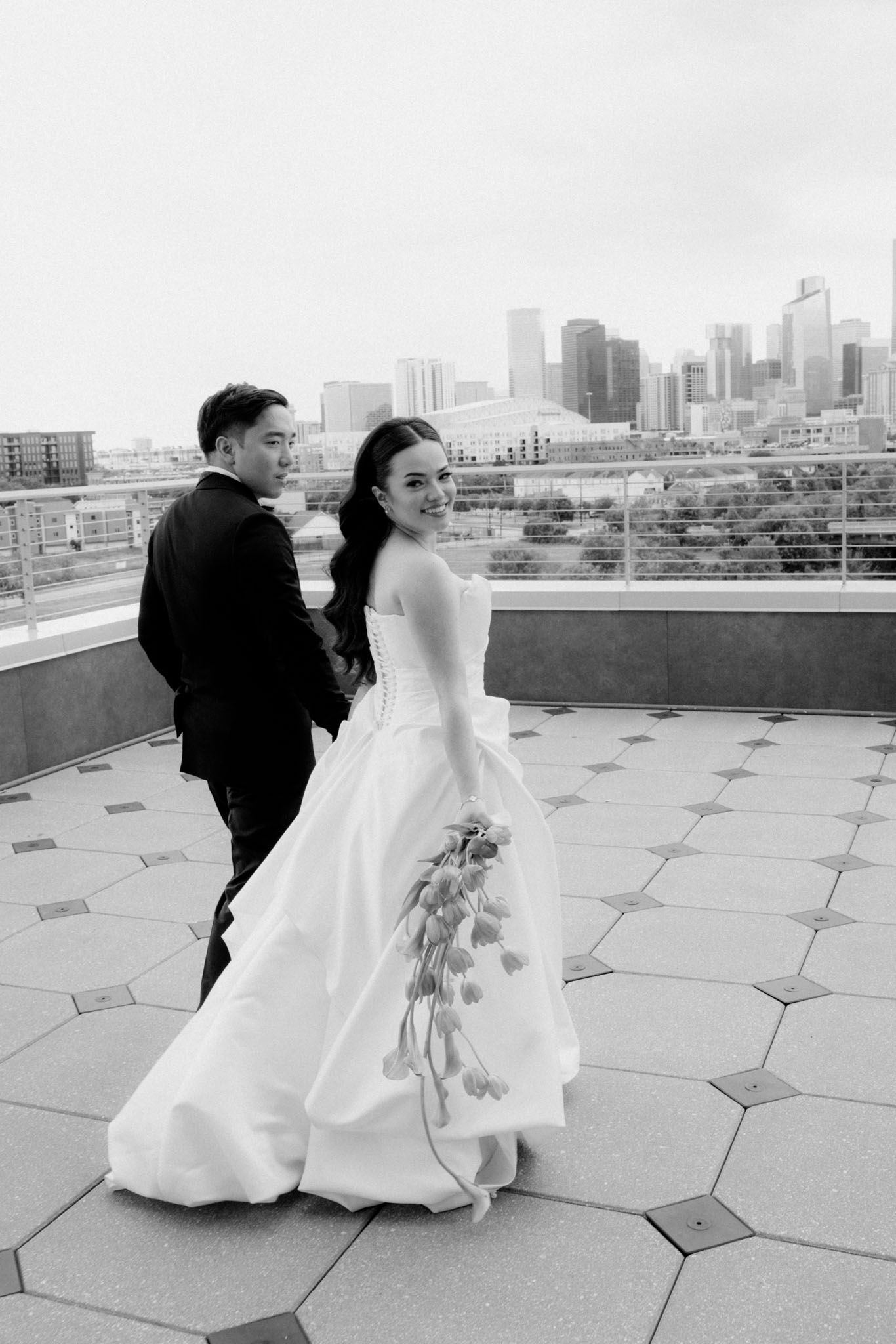 A couple in wedding attire dances together on a rooftop terrace with a city skyline in the background.