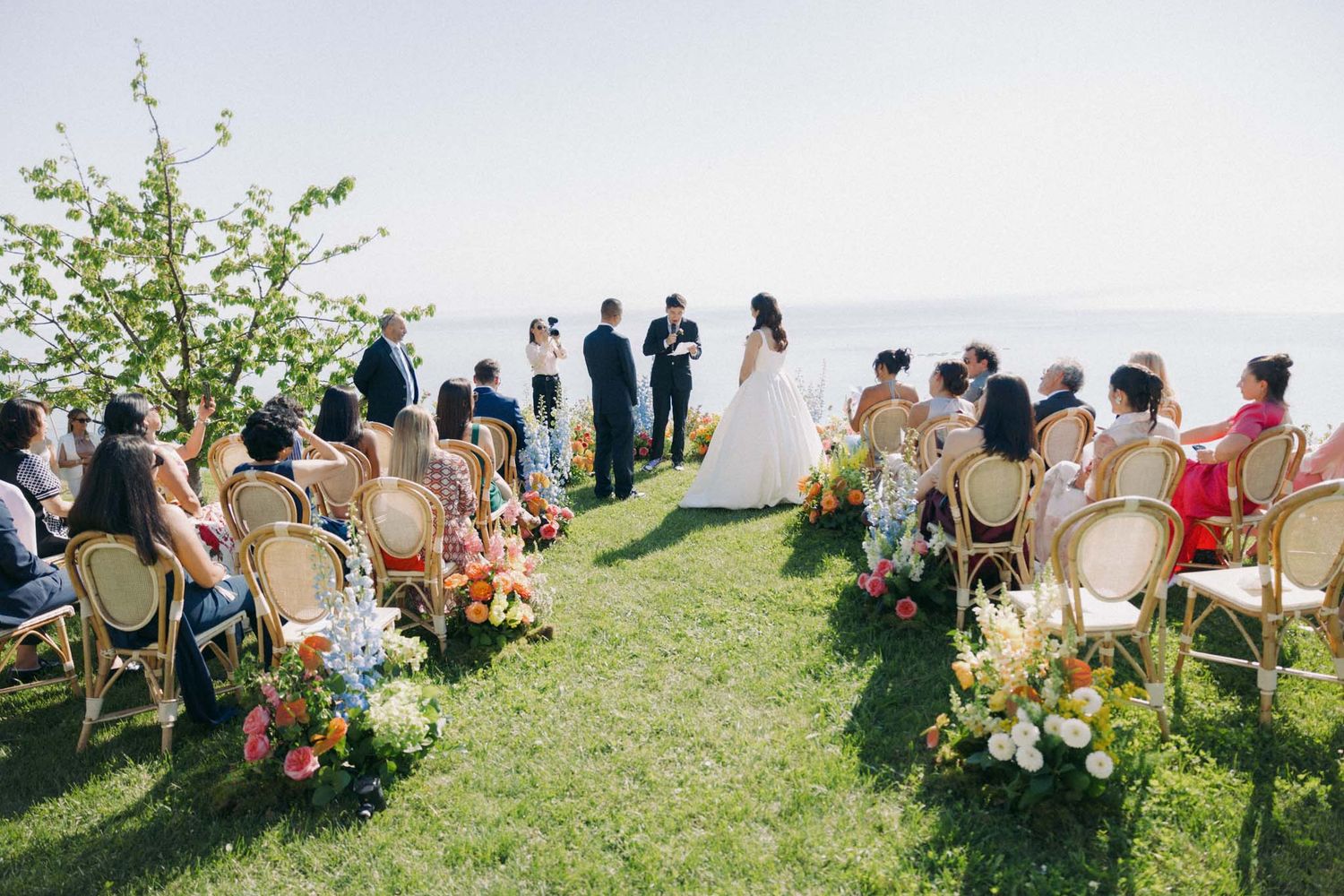 Wedding guests seated in wooden chairs on a grassy lawn during an outdoor ceremony.