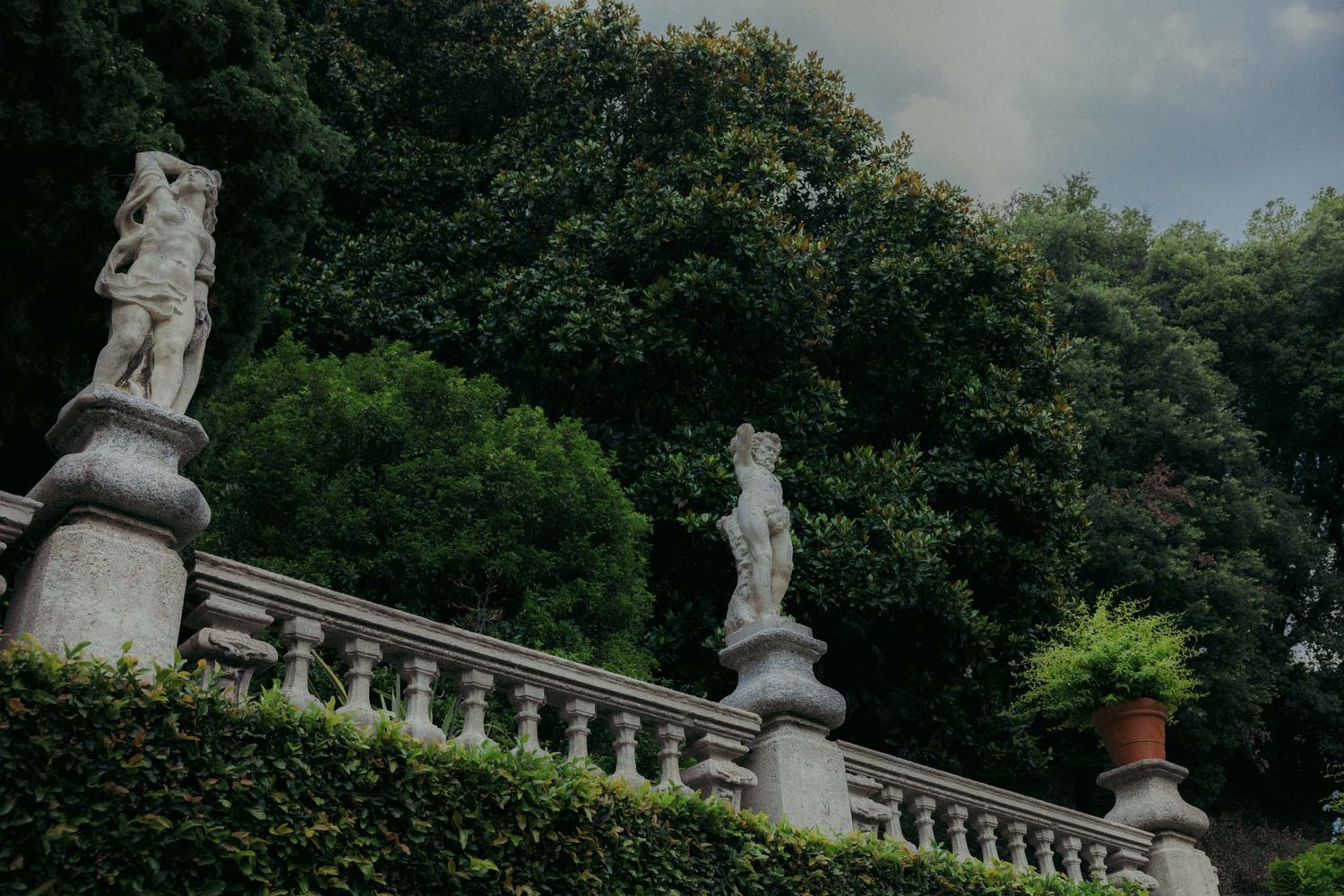 White stone statues and ornate balustrades decorate a formal garden terrace with lush green trees in the background.