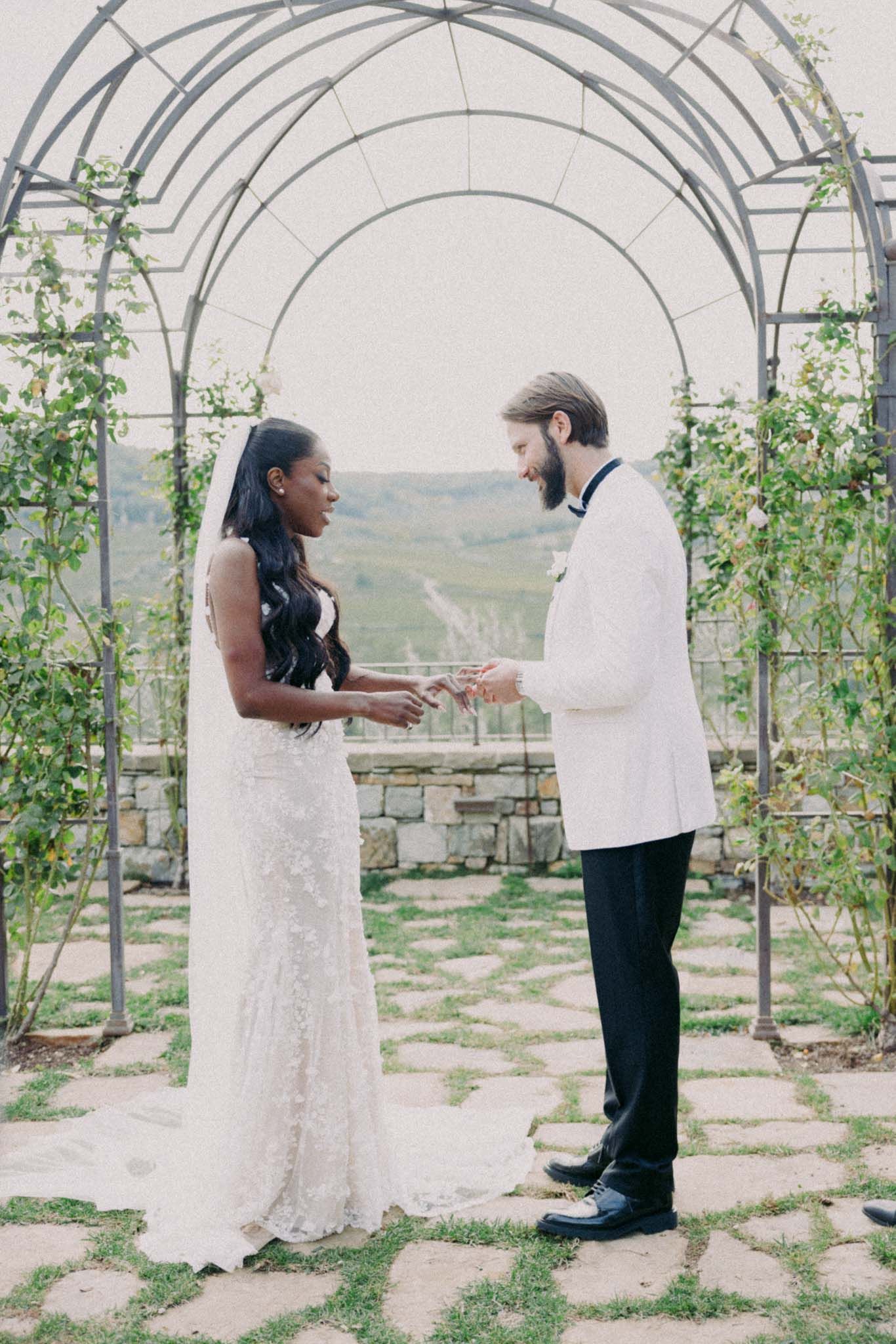 A couple shares a romantic moment under white arches surrounded by climbing vines in a scenic outdoor setting.