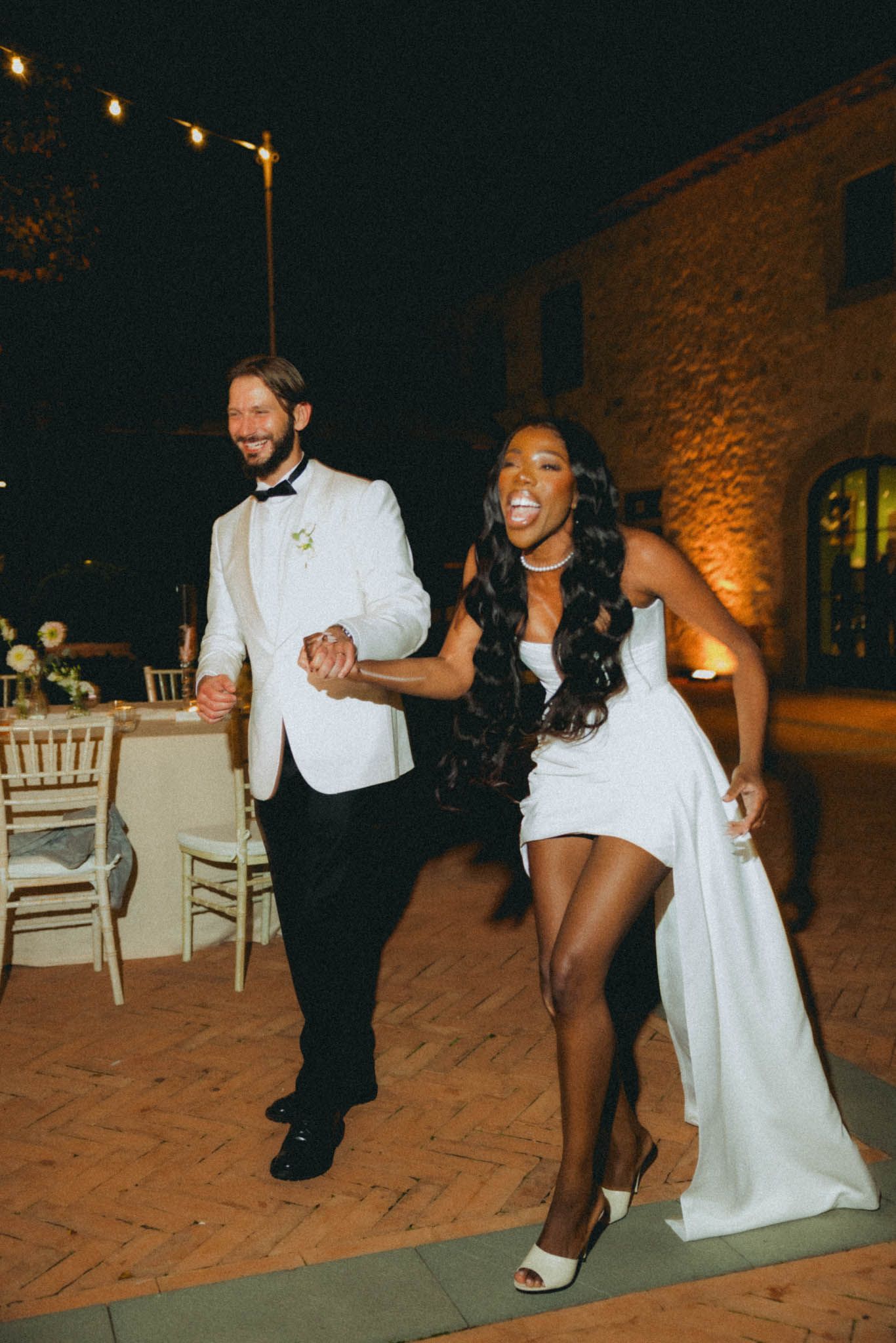 A couple in formal attire dances together at an evening wedding reception on a stone patio.