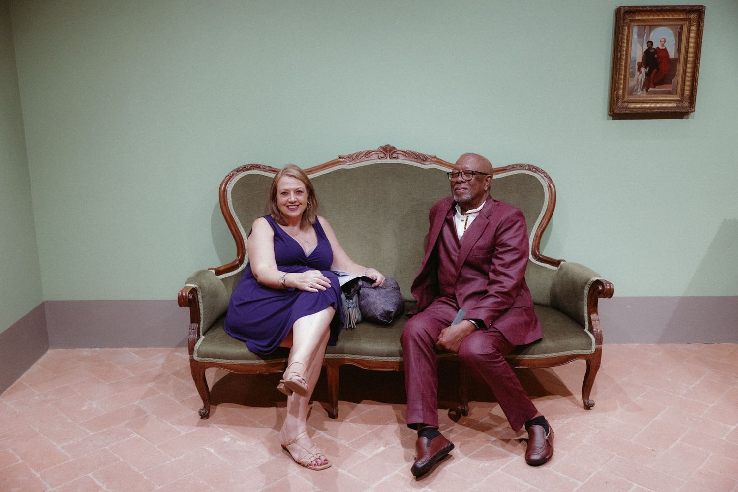 Two people sitting on an antique sofa in a minimalist room with pale green walls and framed artwork.