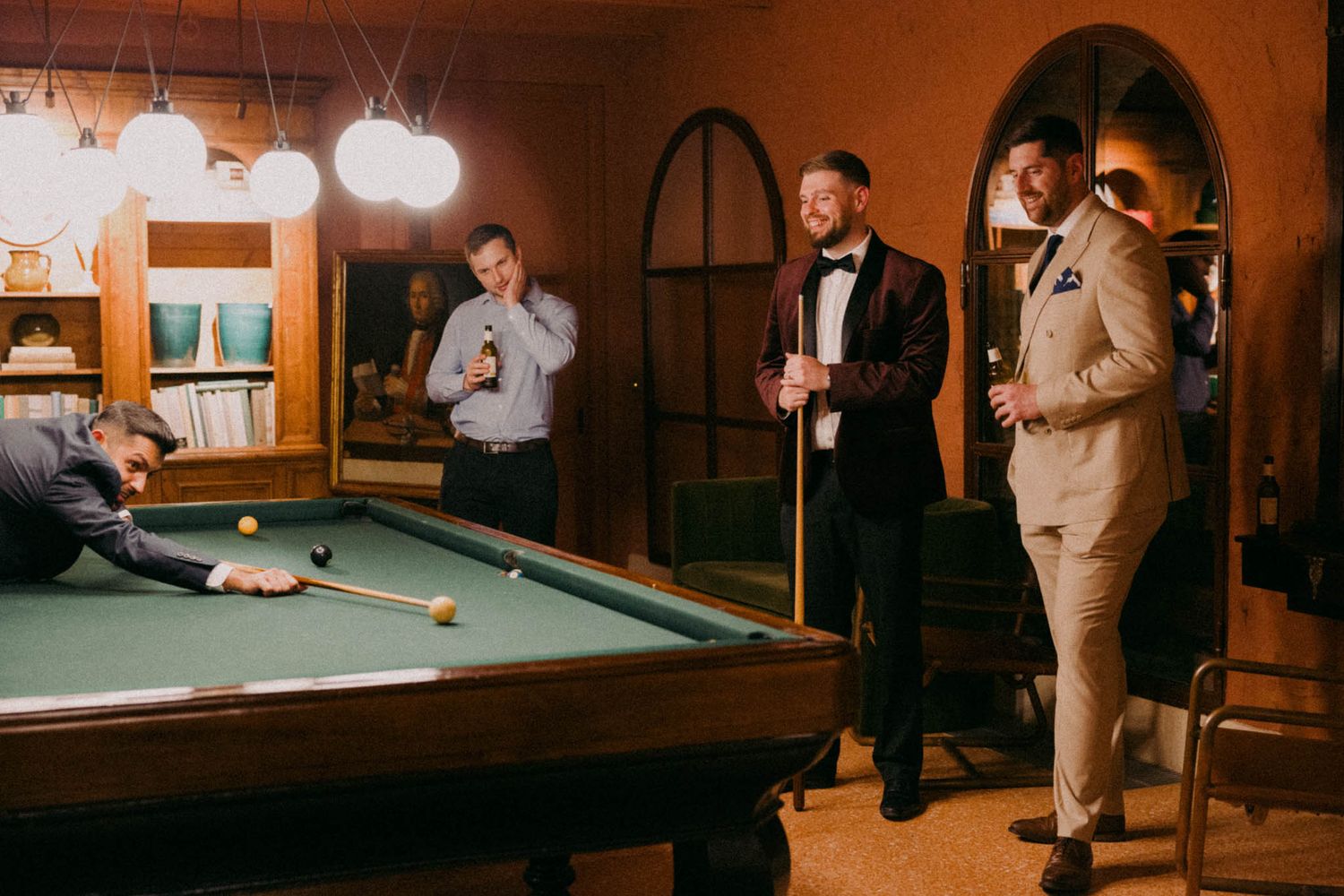 A group plays pool at a wedding reception in a dimly lit room with warm lighting and elegant formal attire.