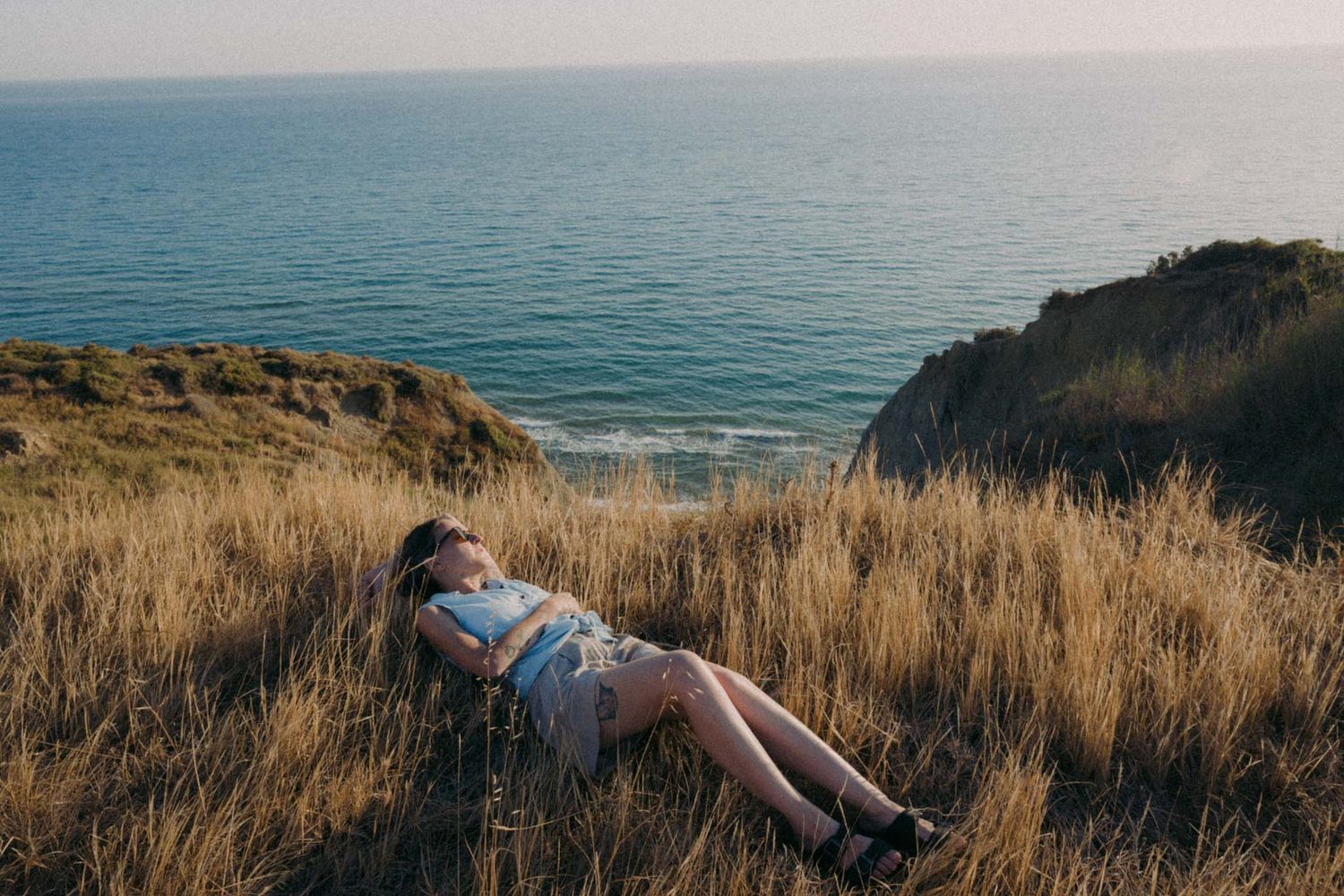 Person lying in tall grass on coastal cliff overlooking turquoise ocean waters at sunset.