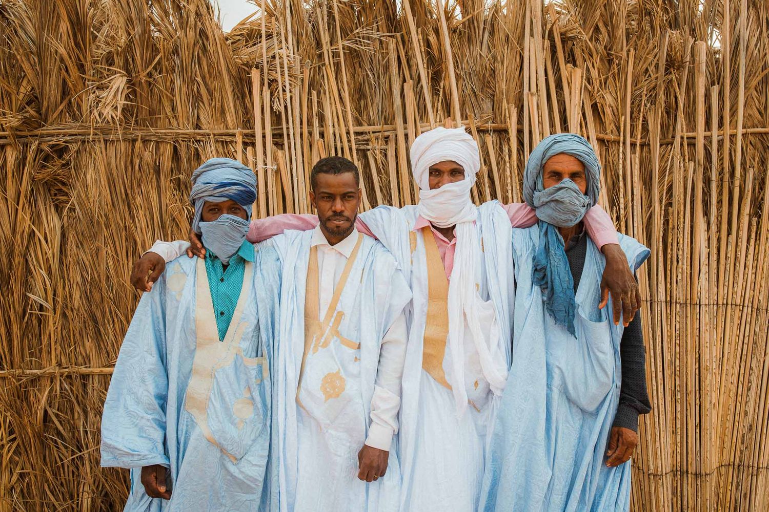 Row of people in traditional white and blue robes stand together against woven wall.