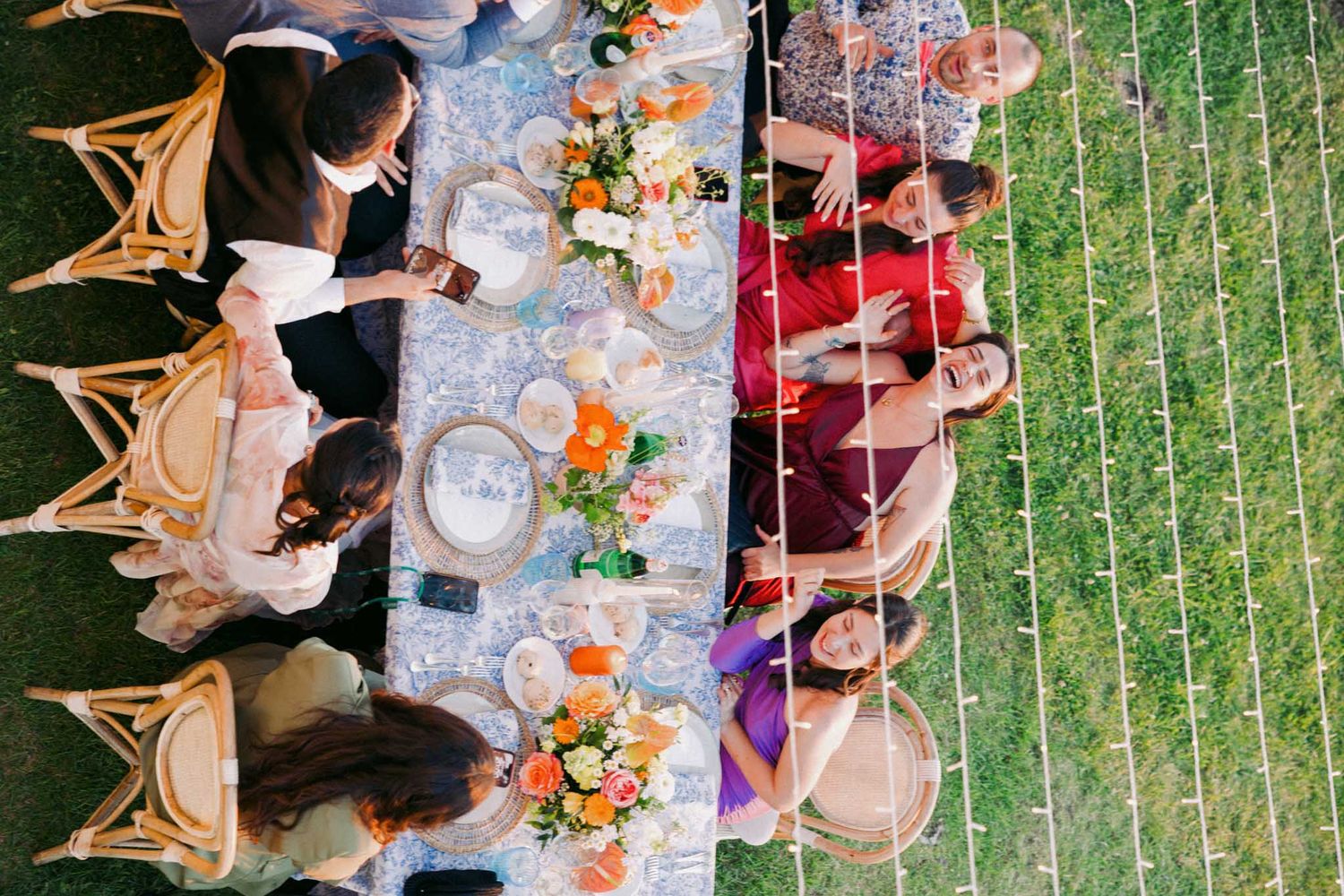 Aerial view of an elegant outdoor dinner party with guests seated at long tables decorated with flowers and glassware.