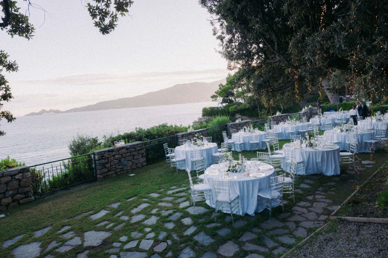 Outdoor wedding reception setup with white tables and chairs arranged on a stone patio overlooking a scenic landscape.
