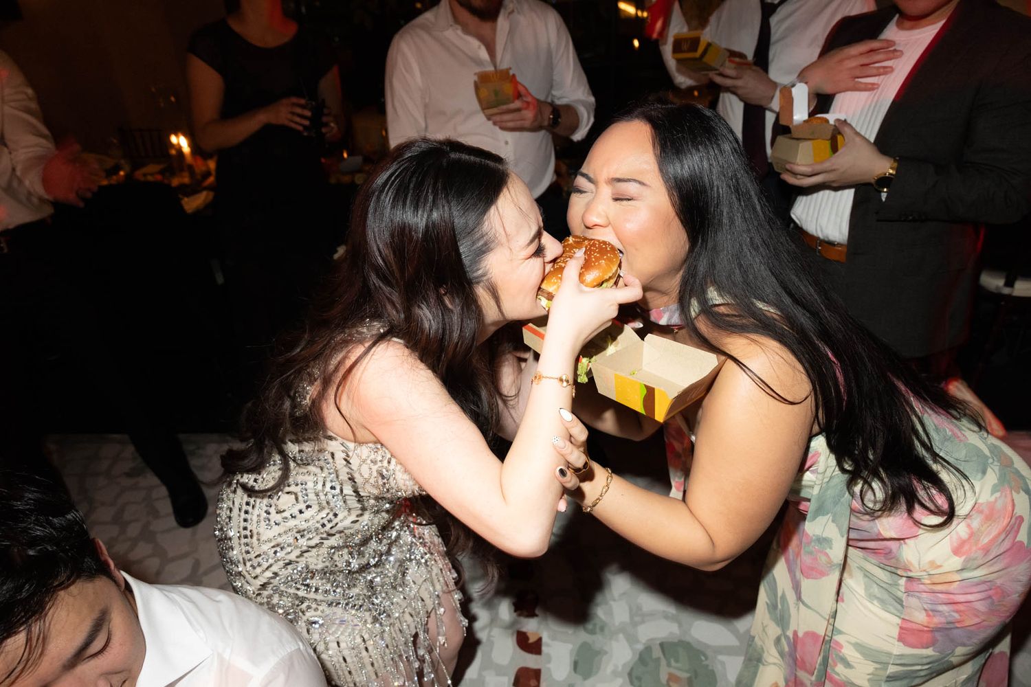Two friends share a laugh while enjoying food and drinks at a lively party gathering.
