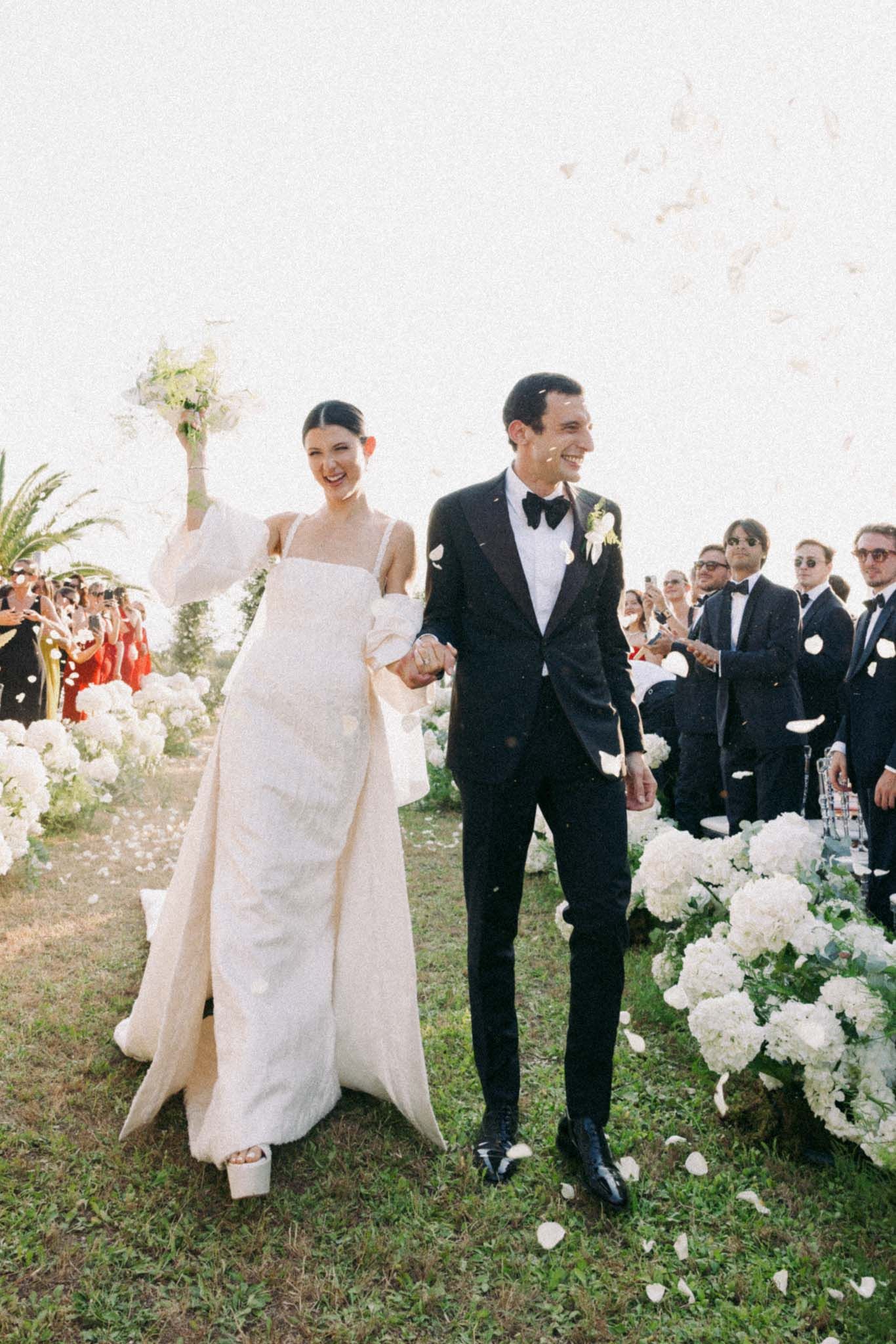 Wedding party walking together outdoors on grassy lawn with white floral arrangements.